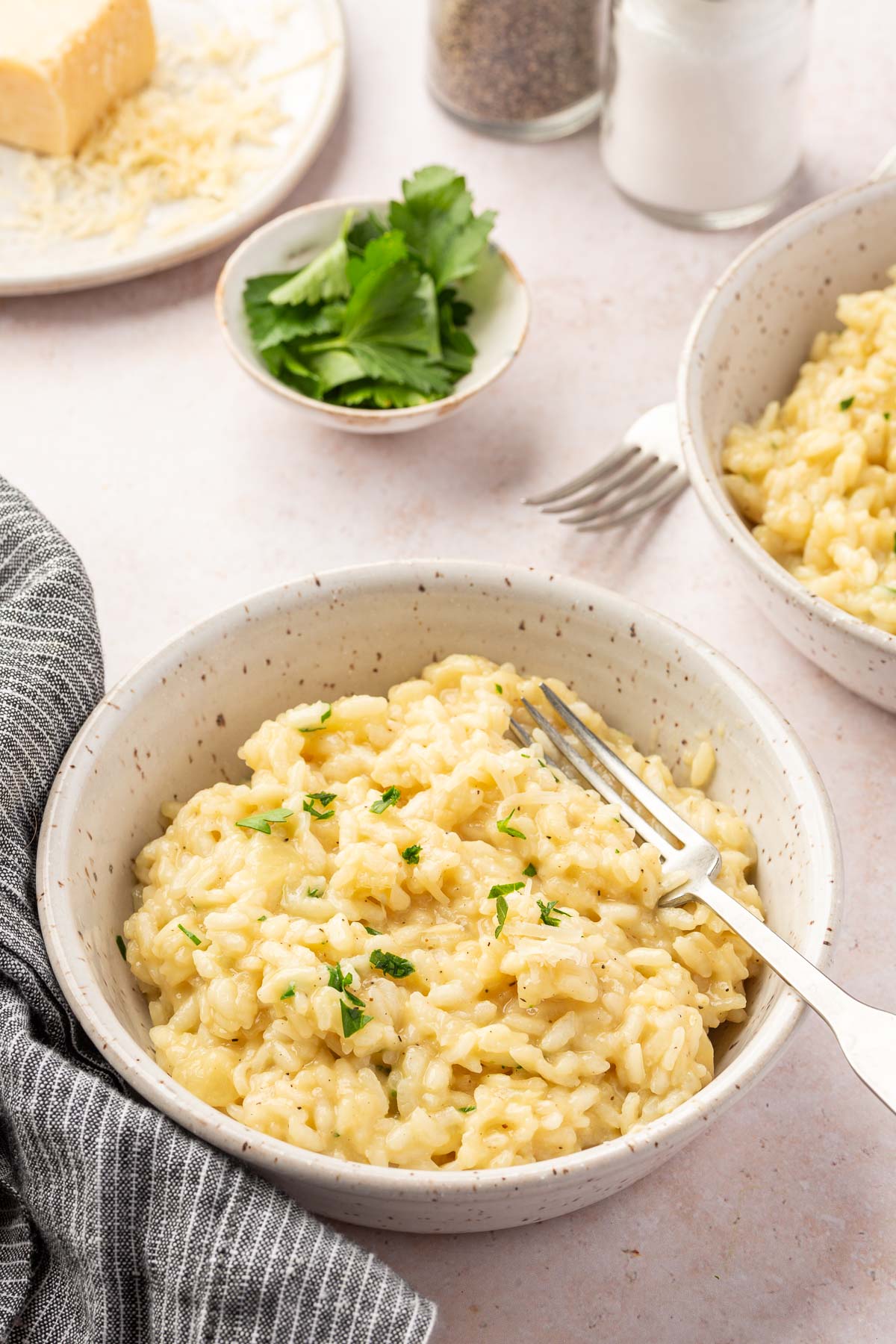 Two bowls of gluten free risotto with forks in them on a table with a small bowl of fresh parsley, a block of parmesan cheese that has been partially shredded, and glass salt and pepper shakers.