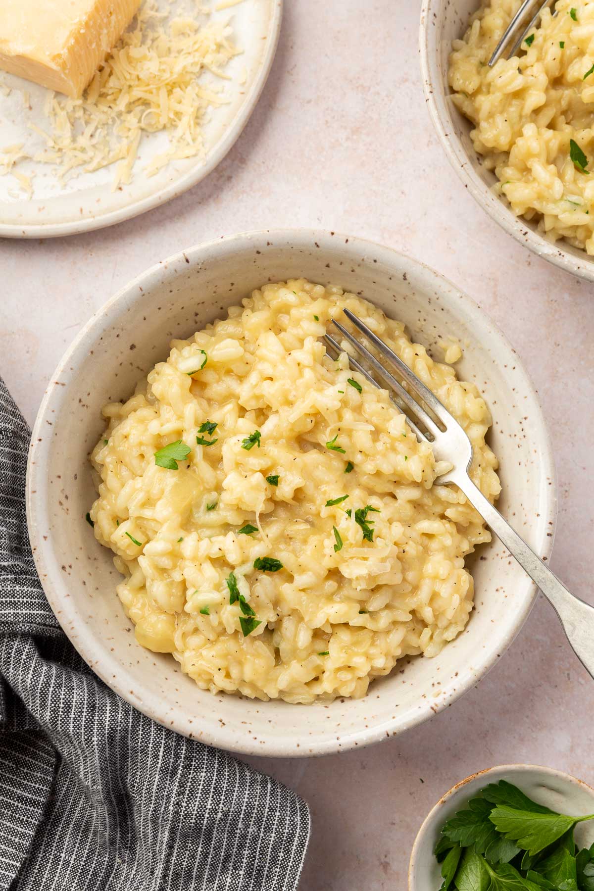 An overhead view of two bowls of gluten free risotto with a bowl of parmesan and a small bowl of fresh parsley.