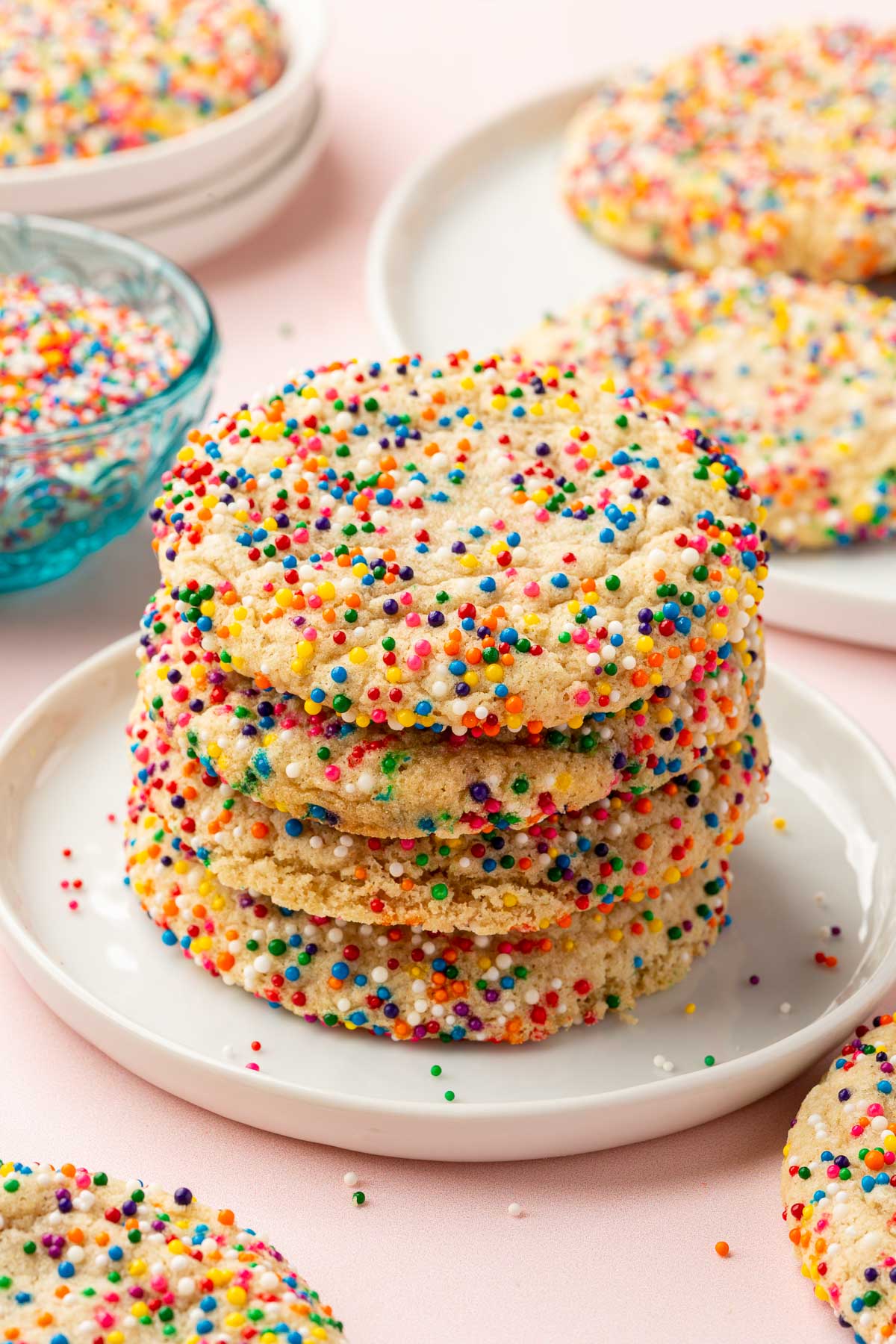 A stack of four gluten-free sprinkle sugar cookies on a dessert plate with additional cookies and sprinkles in the background.