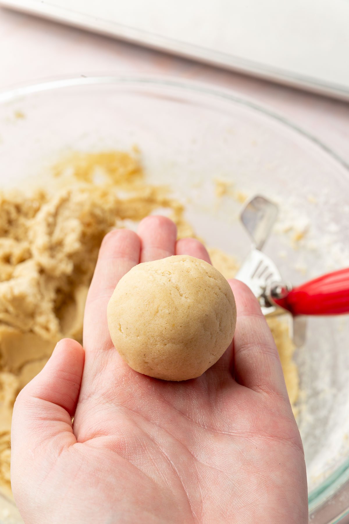 A hand holding a ball of sugar cookie dough over a bowl of additional dough.
