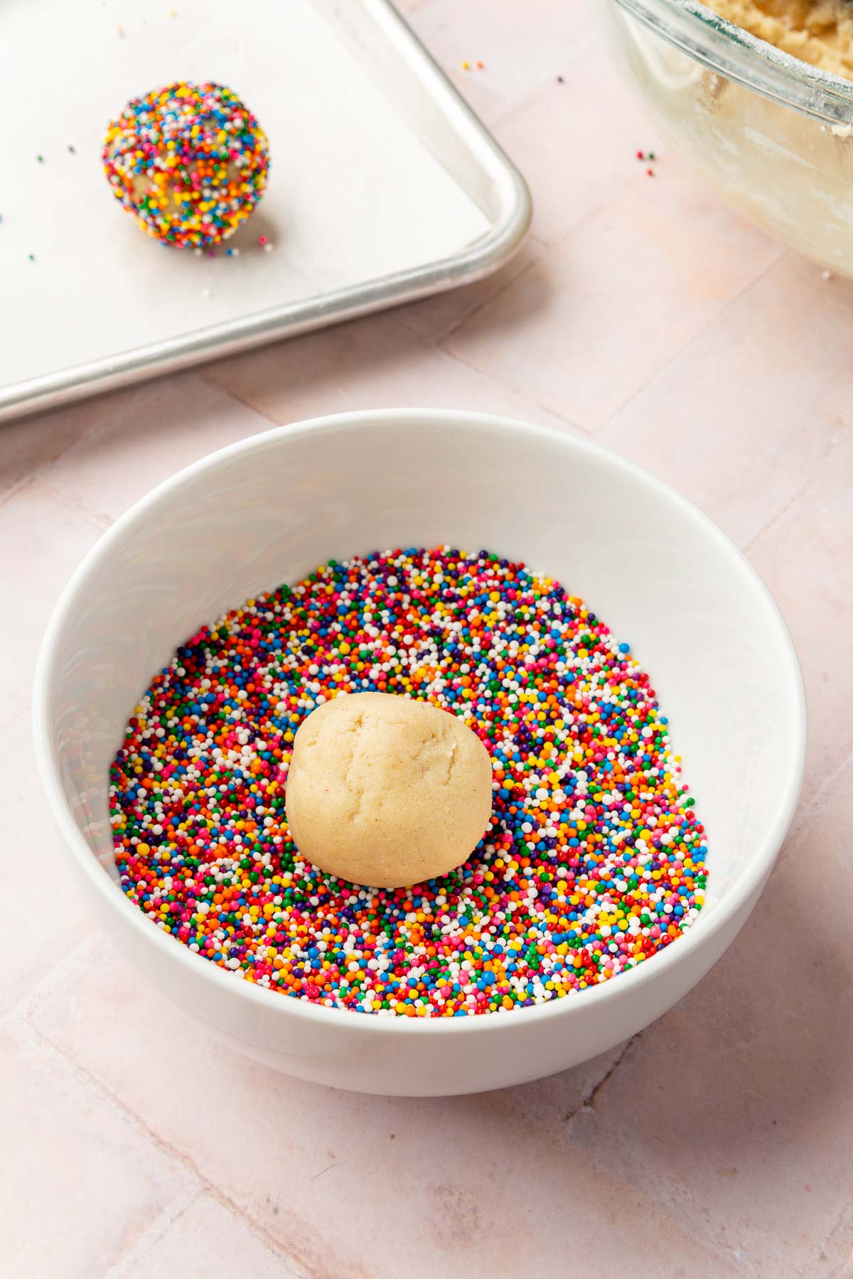 A sugar cookie dough ball in a bowl of non-pareil sprinkles before rolling to make the sprinkles adhere to the dough.