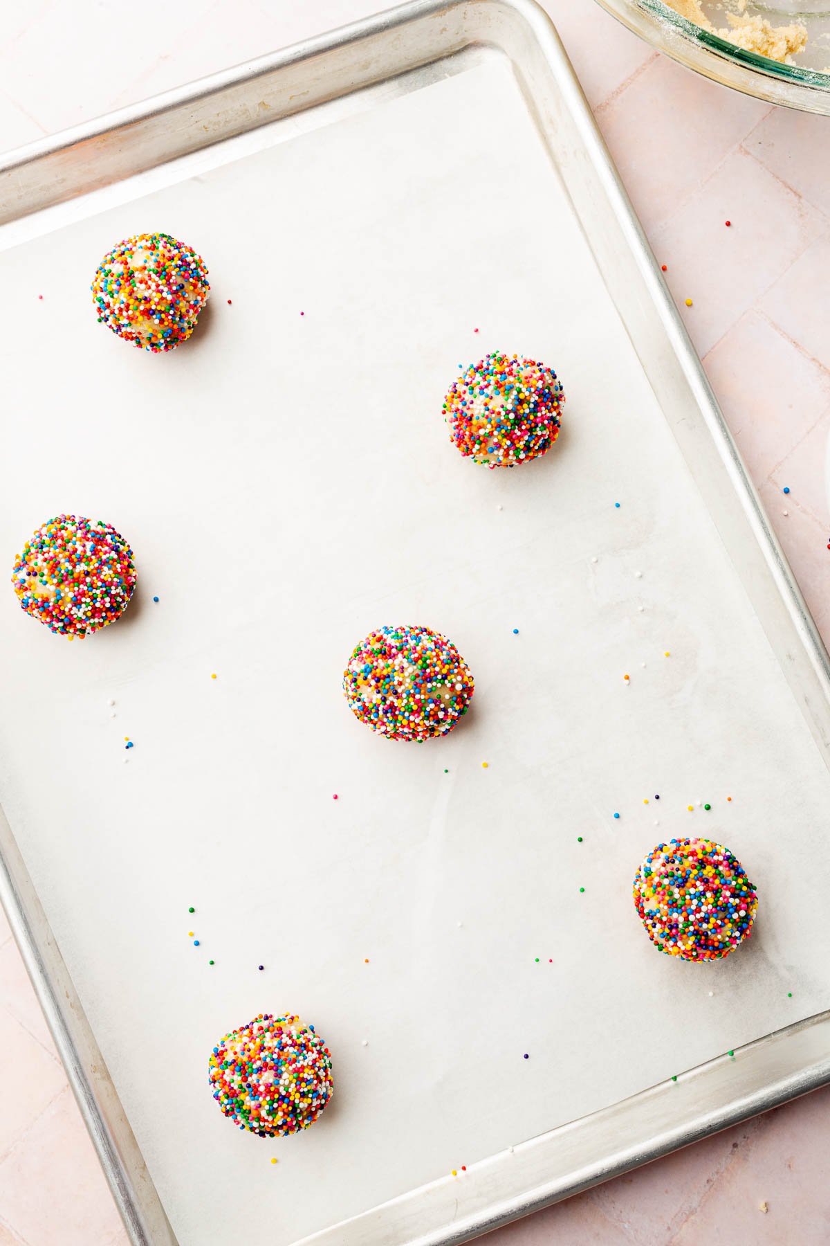 A baking sheet lined with parchment paper and topped with six gluten free sprinkle sugar cookie dough balls before baking in the oven.