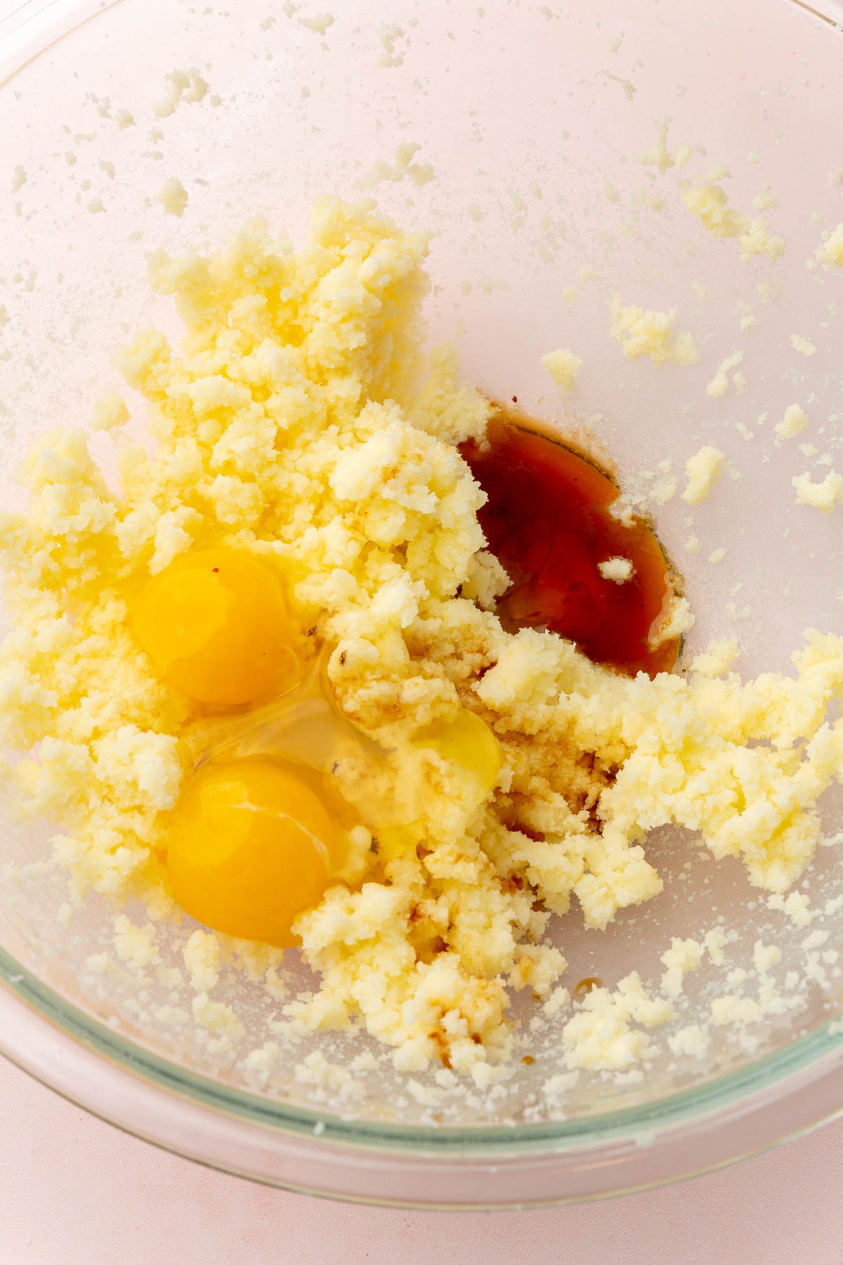 A glass mixing bowl filled with creamed butter and sugar topped with two eggs and vanilla extract before mixing together.
