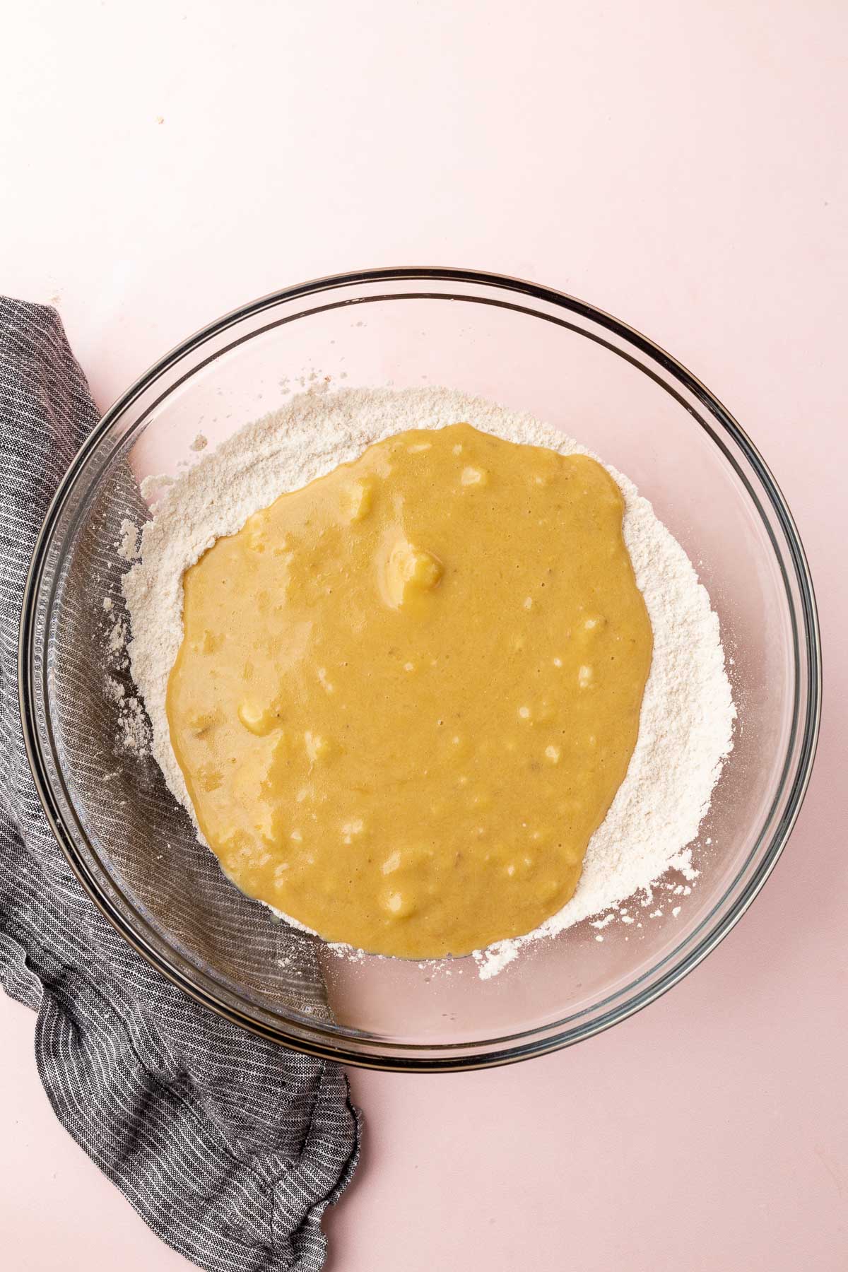 A glass mixing bowl with gluten-free flour topped with a mashed banana, oil and sugar mixture to make banana muffins.