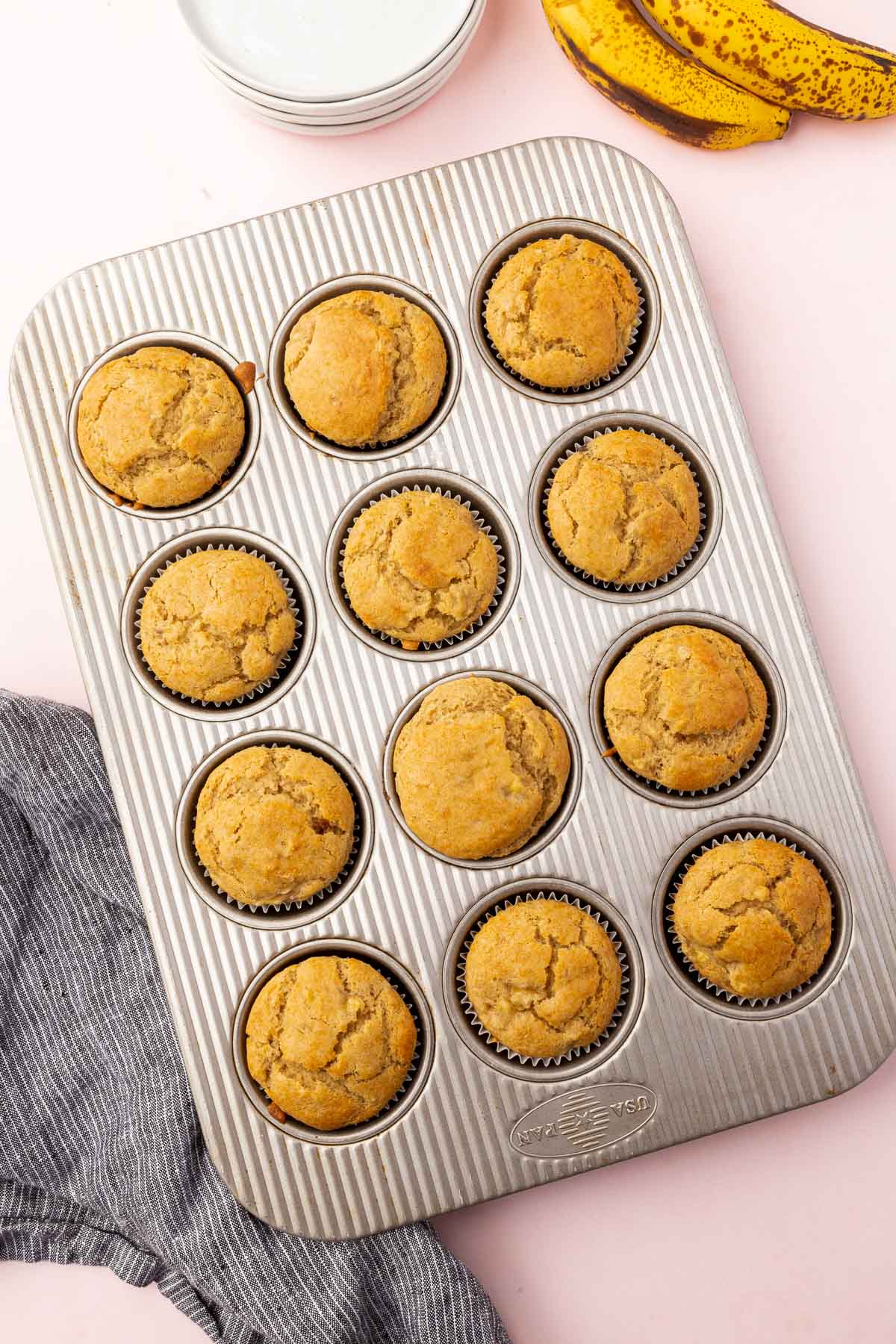 A doze gluten-free banana muffins in a silver muffin tin with ripe bananas to the side.