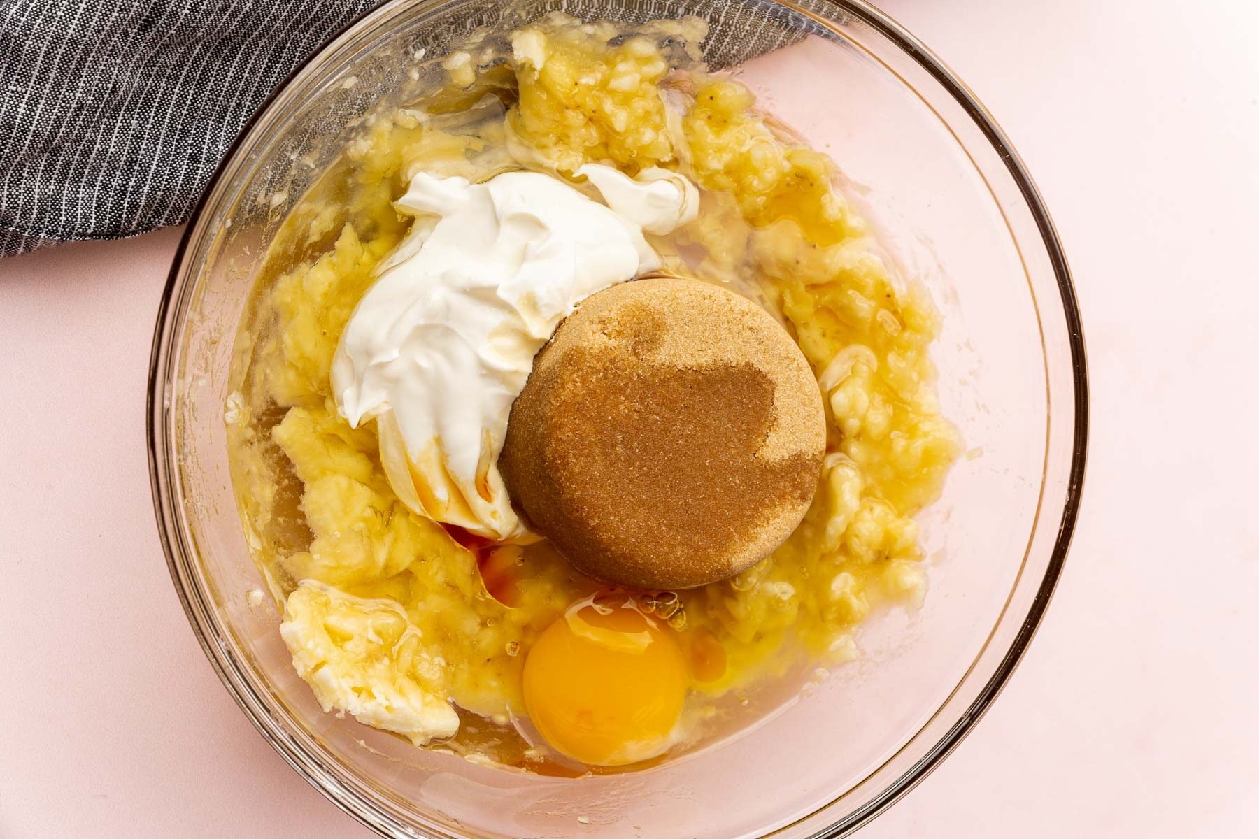 A glass mixing bowl with mashed banana topped with brown sugar, sour cream, oil, egg, and vanilla before whisking together.