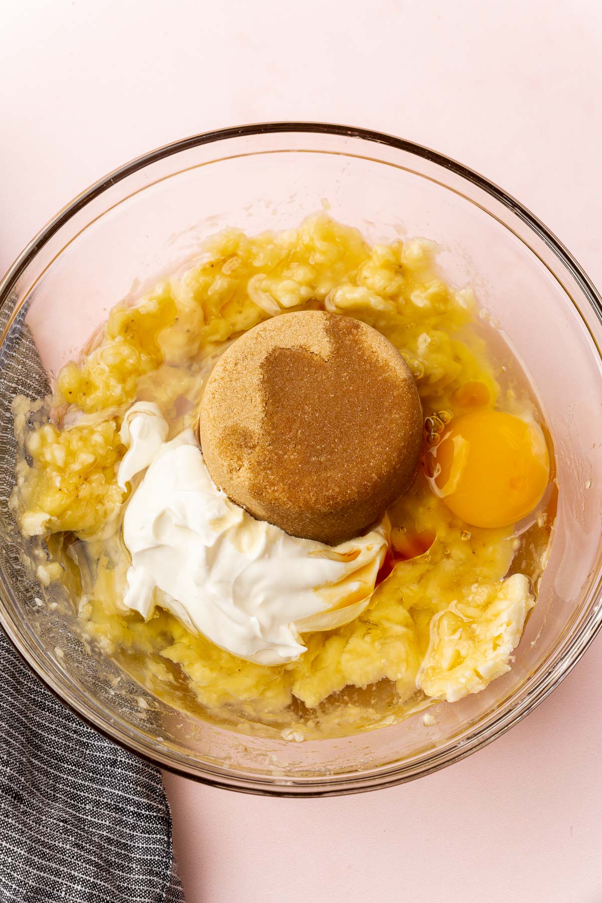 A glass mixing bowl with mashed banana topped with brown sugar, sour cream, oil, egg, and vanilla before whisking together.