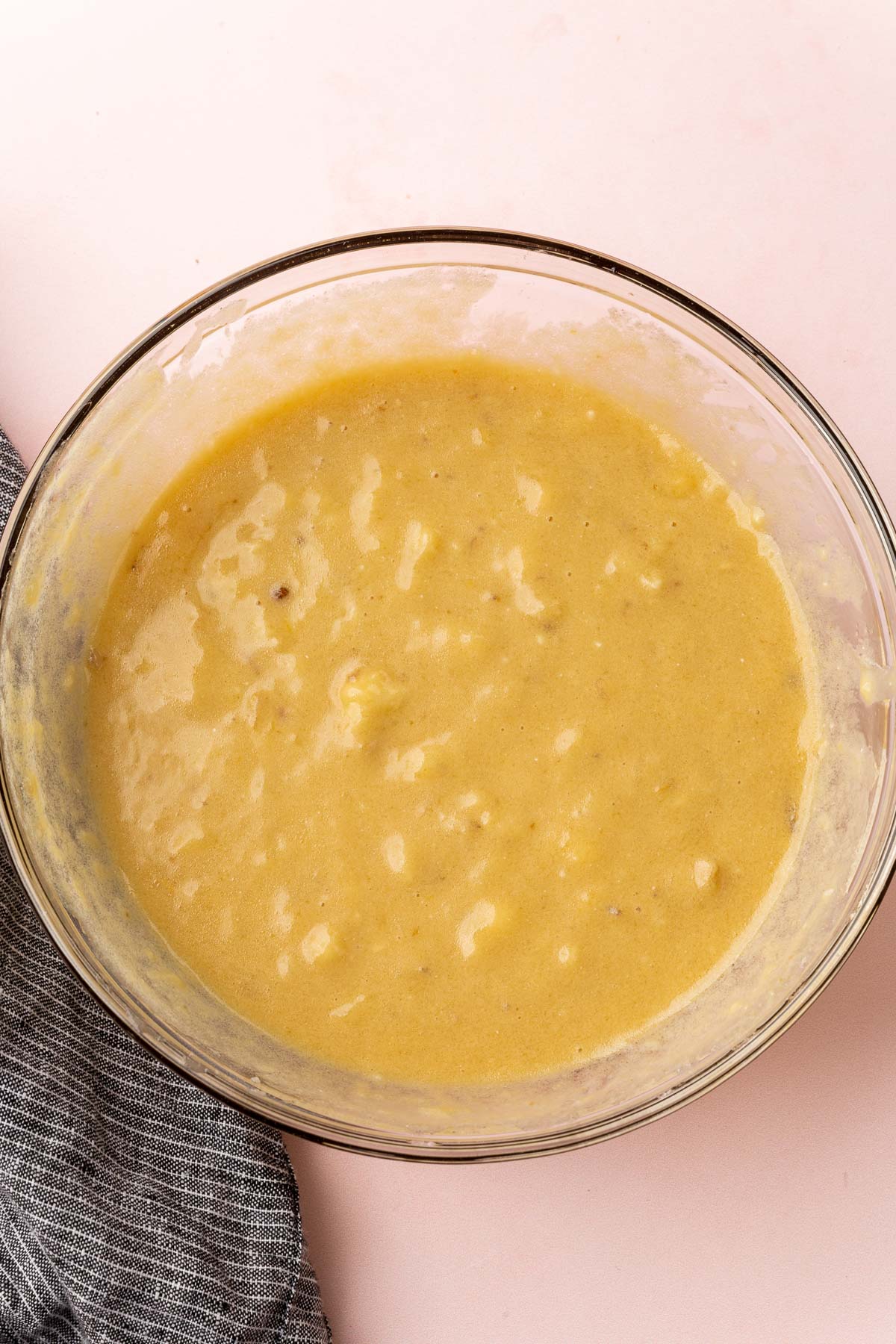 A glass mixing bowl with a mashed banana, brown sugar, oil, egg and vanilla extract mixture in it to make banana muffins.