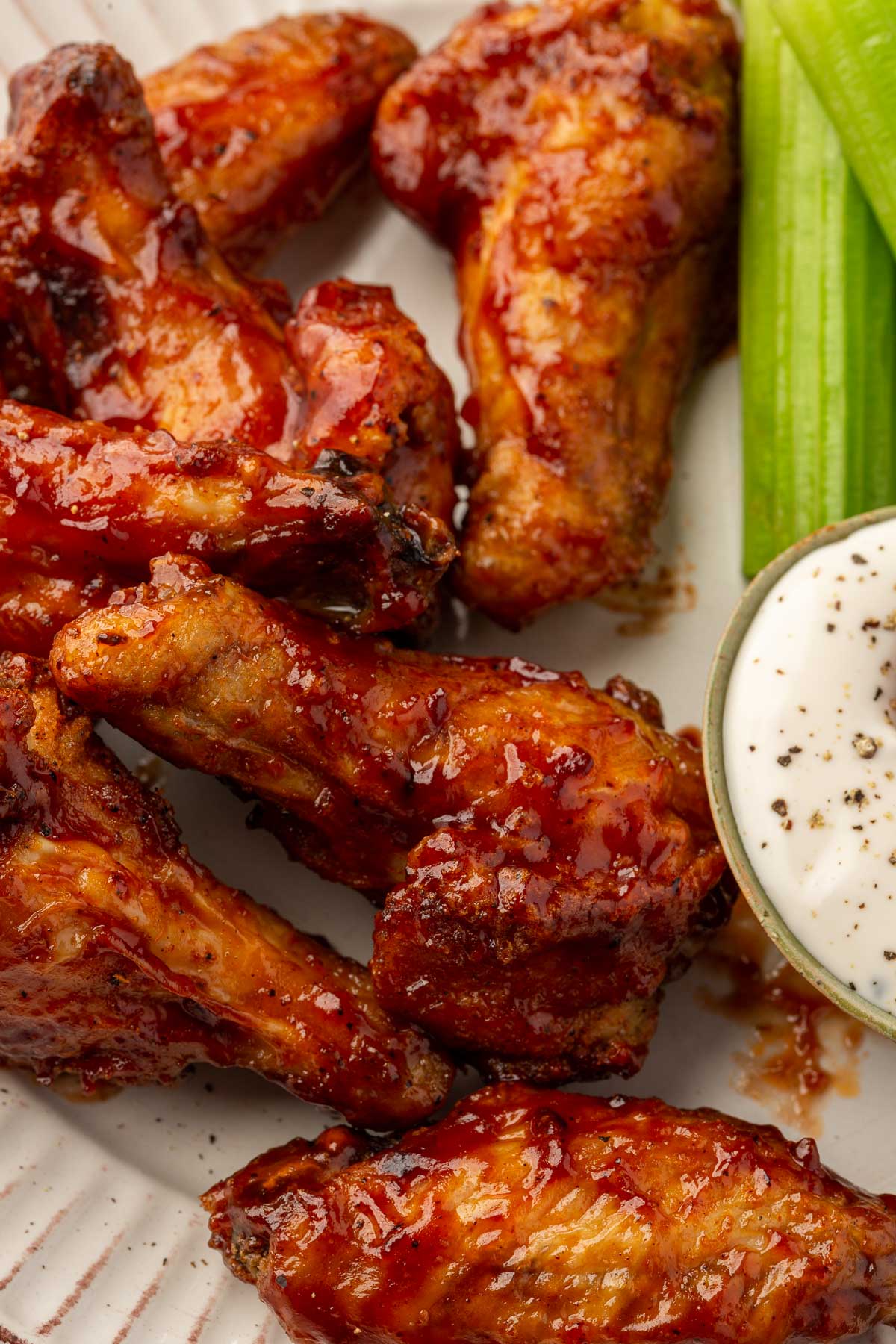 A close up of gluten free chicken wings tossed in BBQ sauce on a plate with pieces of celery and a small bowl of bleu cheese dressing on the side.