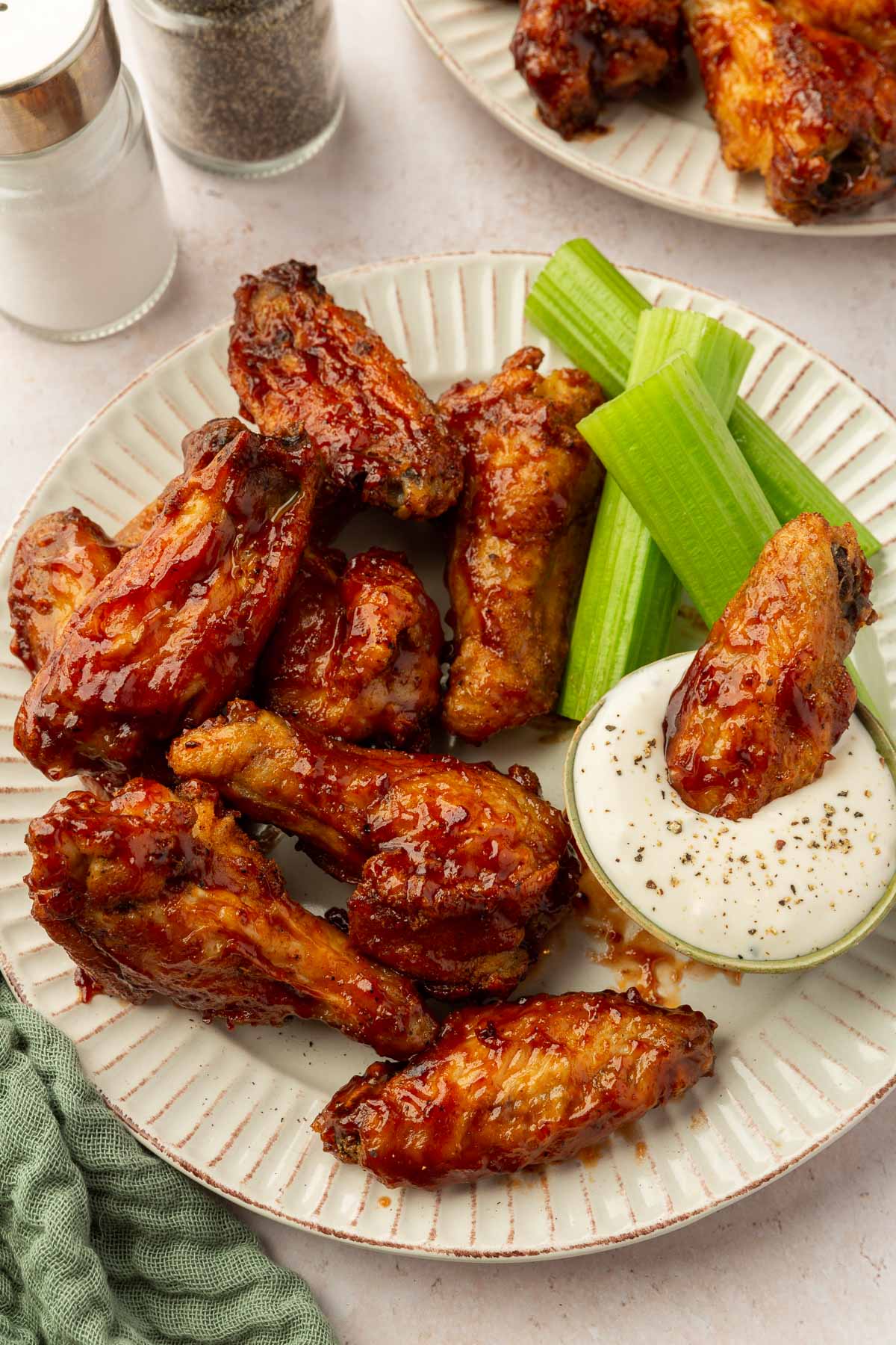 A heaping pile of BBQ chicken wings on a plate with a few pieces of celery and a small bowl of blue cheese dressing with salt and pepper shakers on the side.