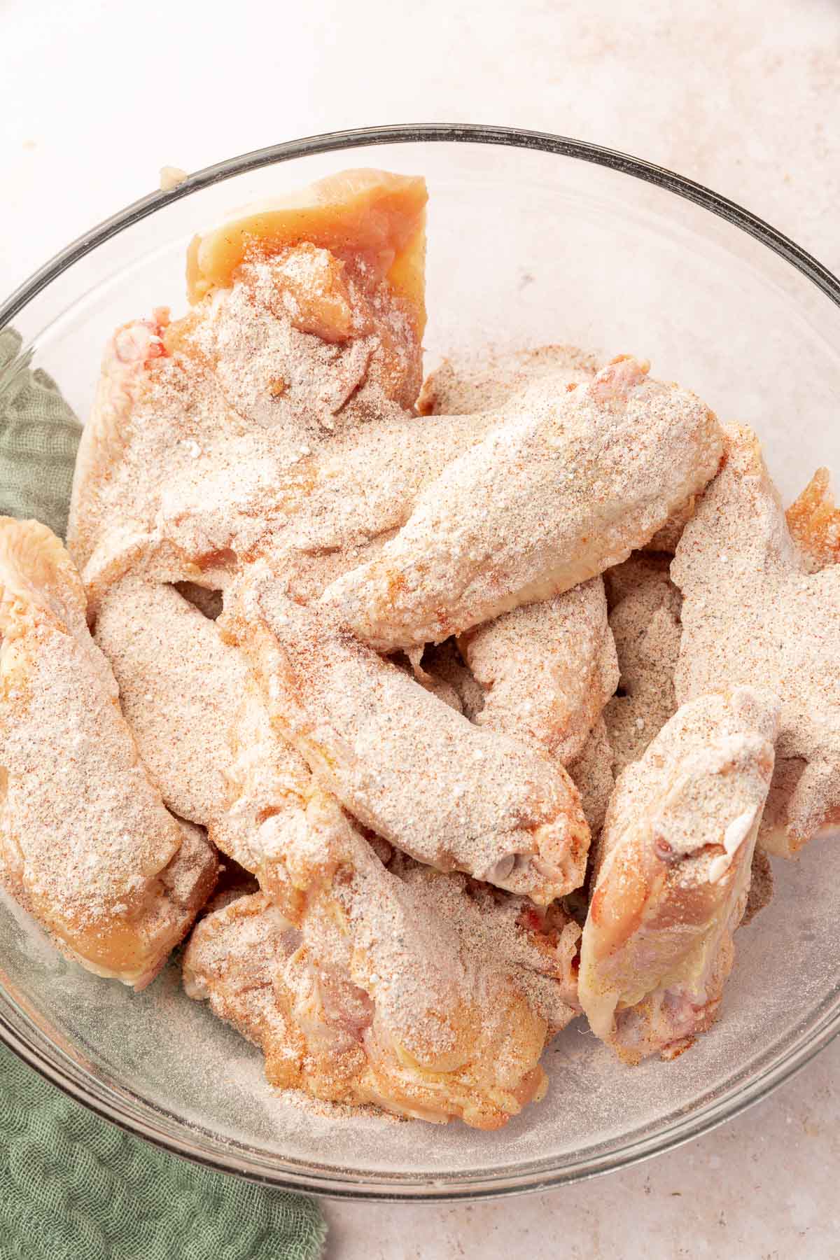A glass mixing bowl with raw chicken wings topped with a mixture of baking powder and spices.