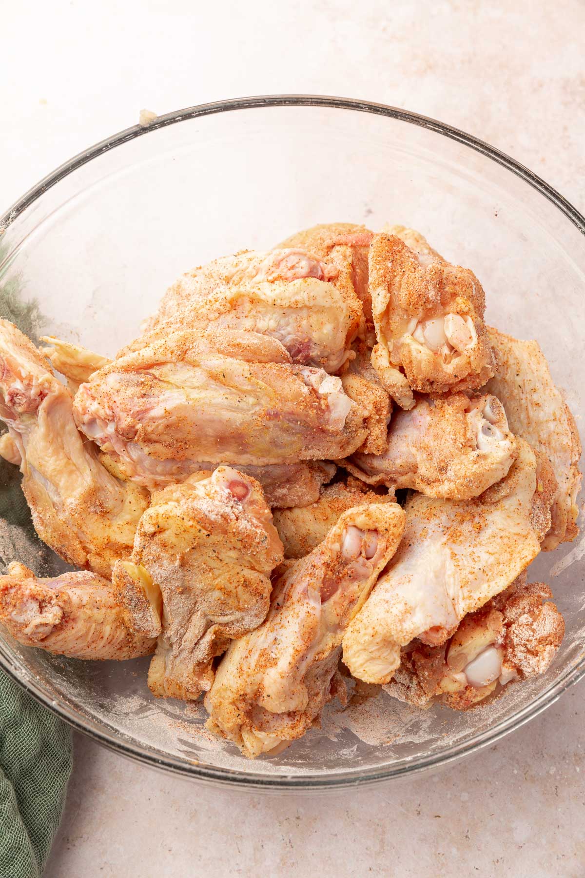 A glass mixing bowl with raw chicken wings tossed in baking pwoder and spices.