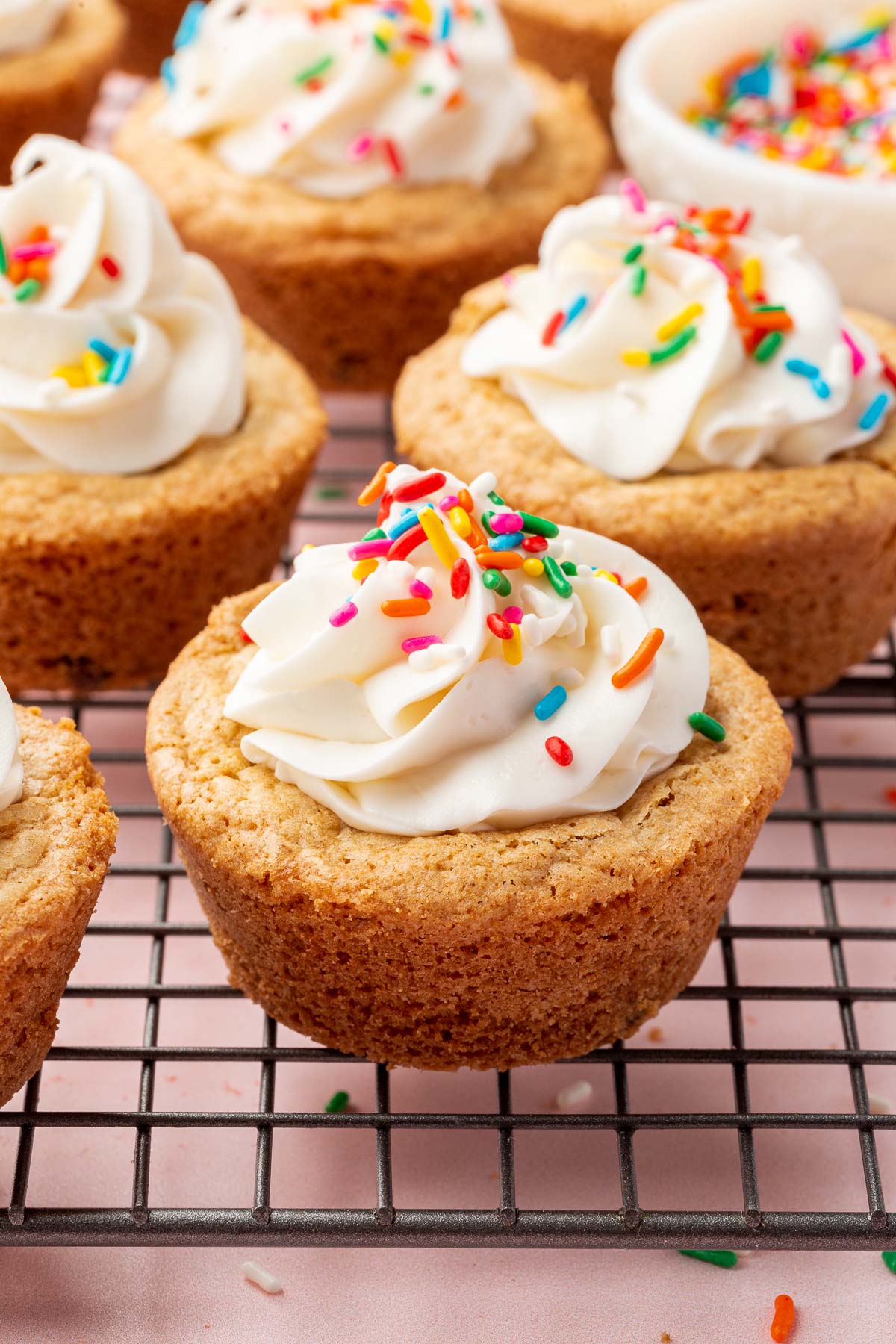 Multiple gluten-free cookie cups topped with vanilla frosting and rainbow sprinkles on a wire cooling rack.
