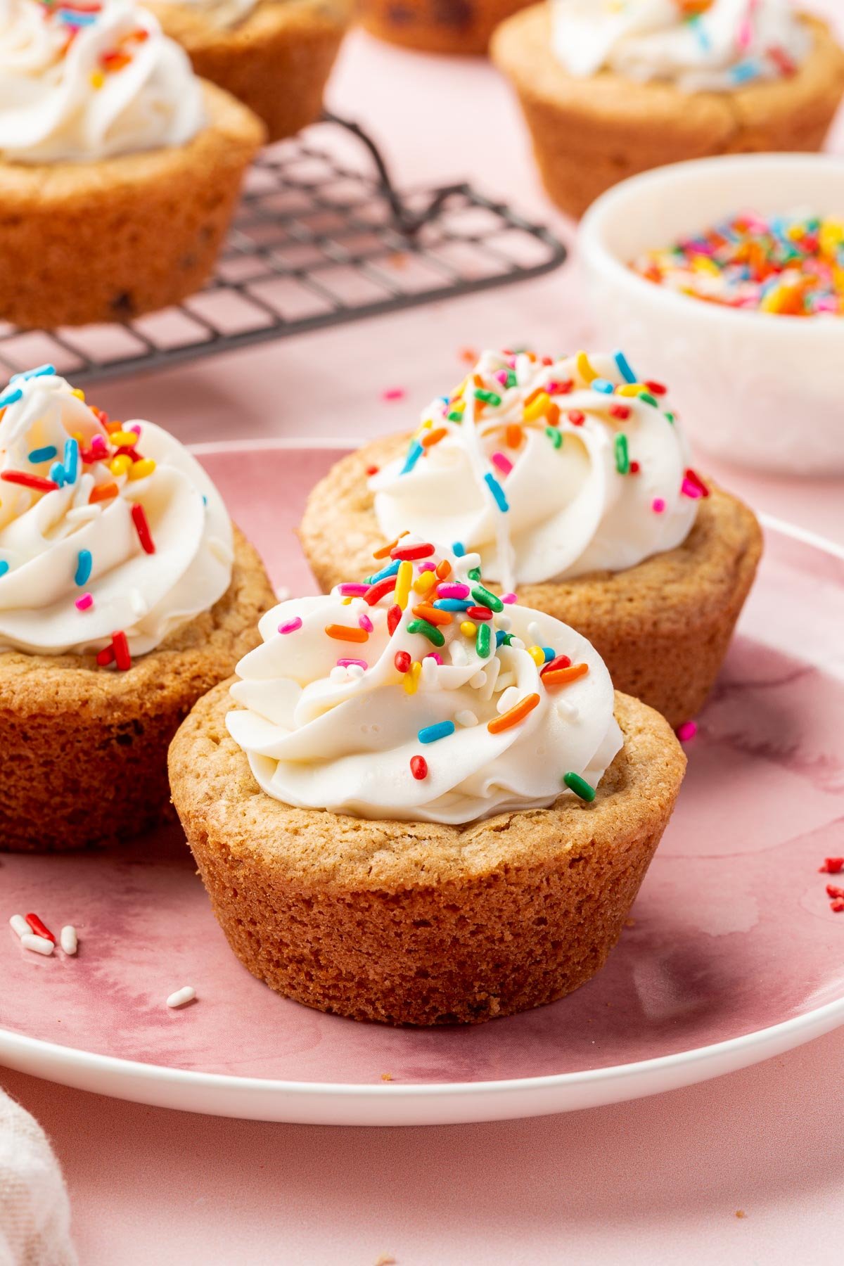 Three gluten-free cookie cups topped with vanilla frosting and rainbow sprinkles on a pink dessert plate with additional cookie cups in the background.