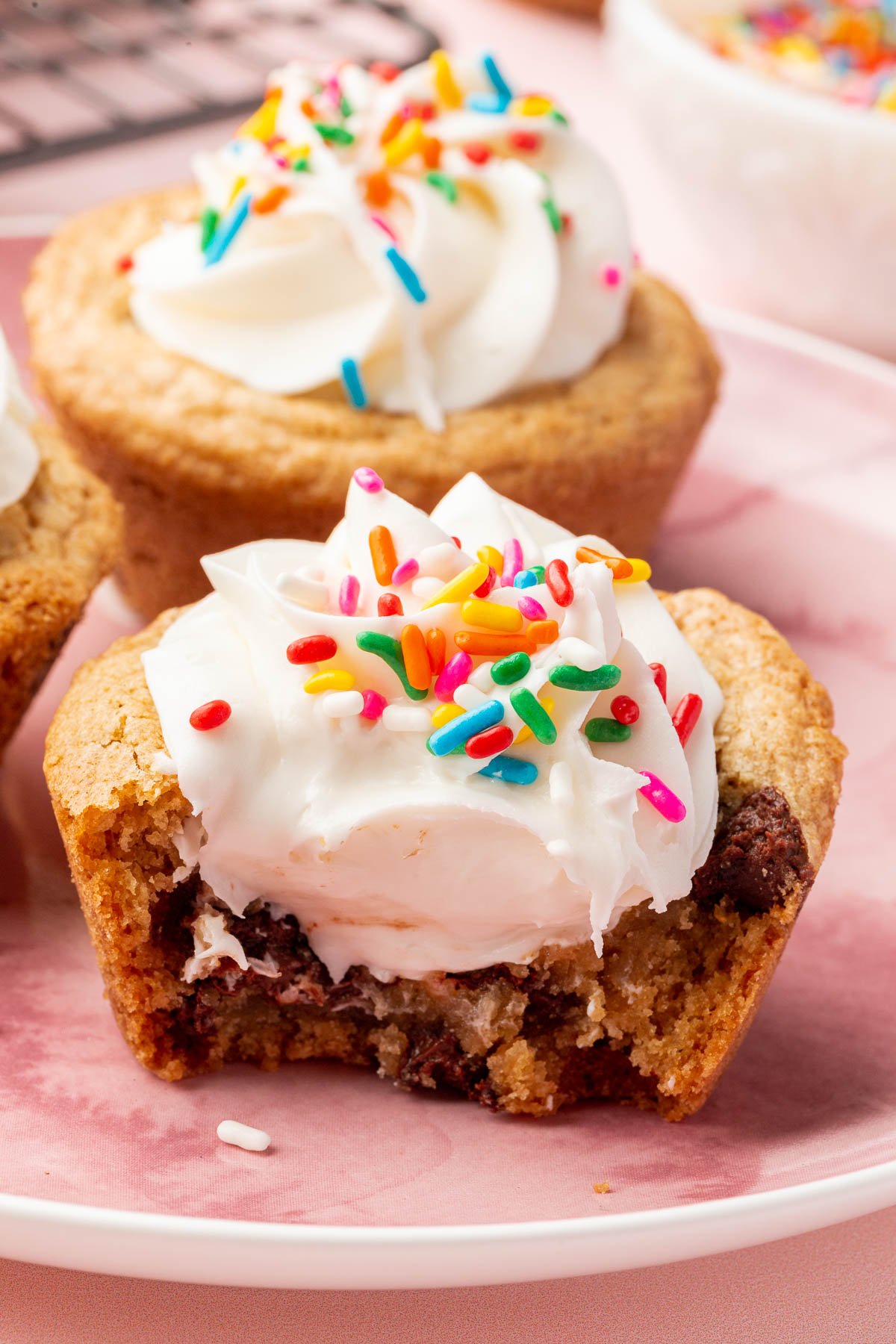 A chocolate chip cookie cup filled with vanilla frosting and topped with rainbow sprinkles that has a bite taken out of it.
