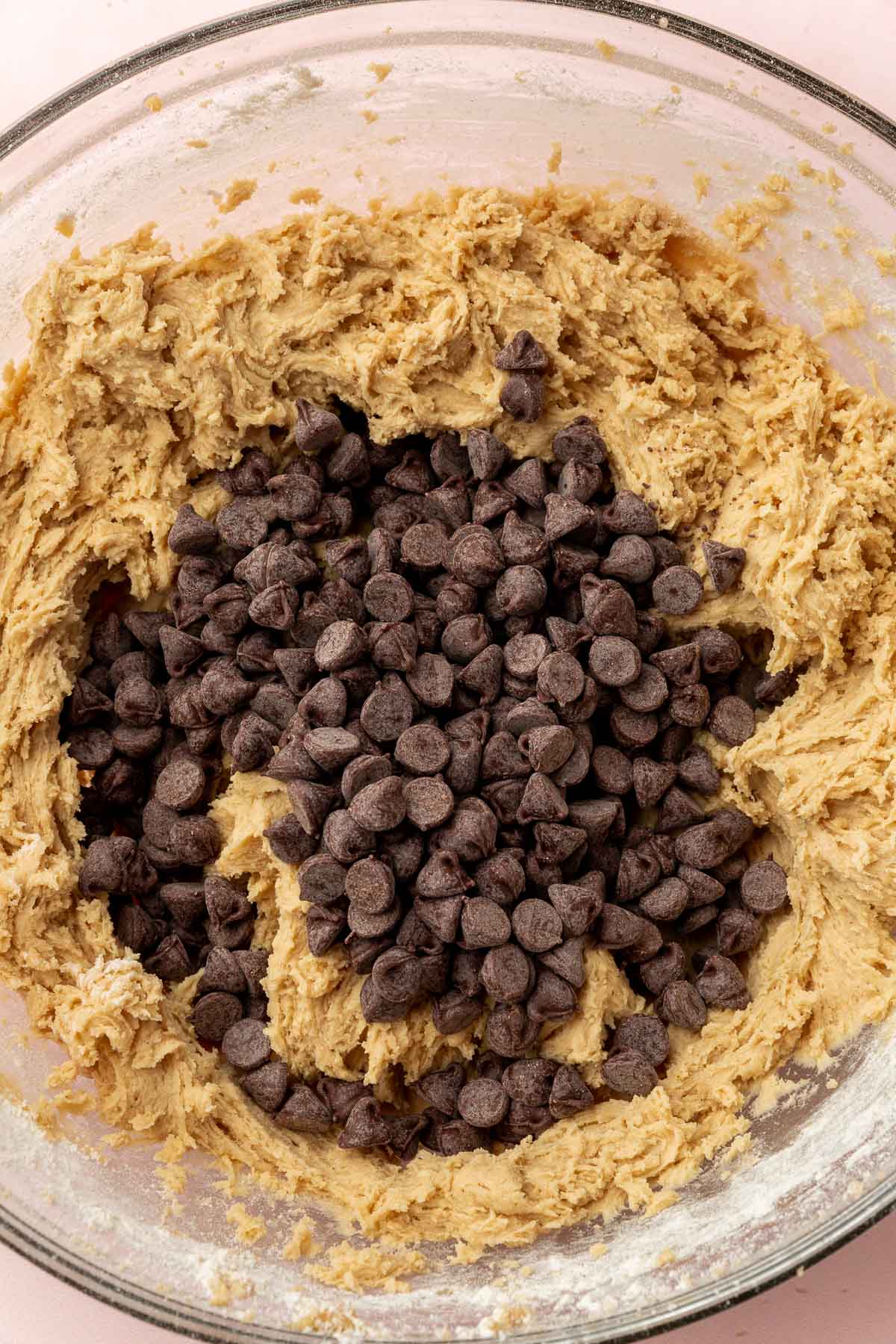 A glass mixing bowl with gluten-free cookie dough topped with a pile of semi-sweet chocolate chips before mixing together.