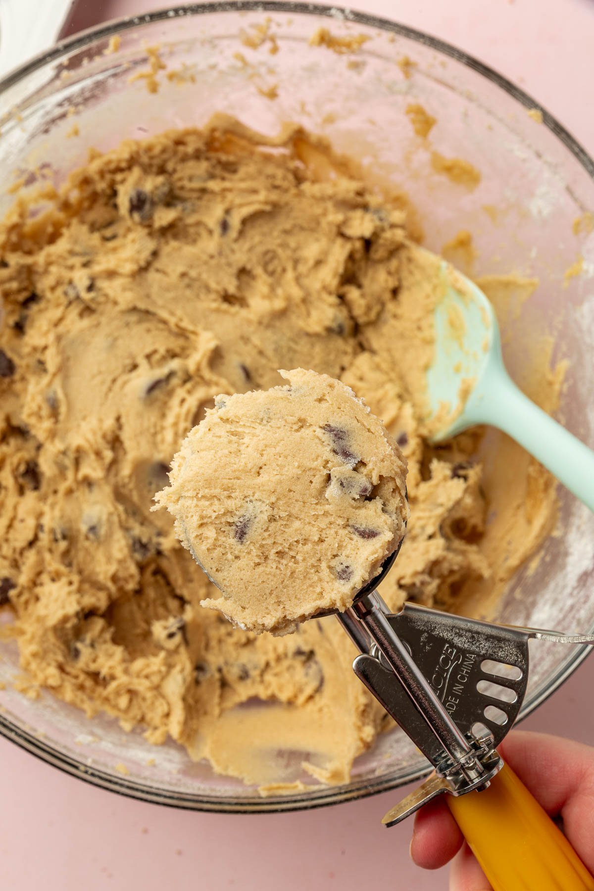 A portion scoop filled with gluten-free chocolate chip cookie dough over a glass mixing bowl of additional cookie dough.