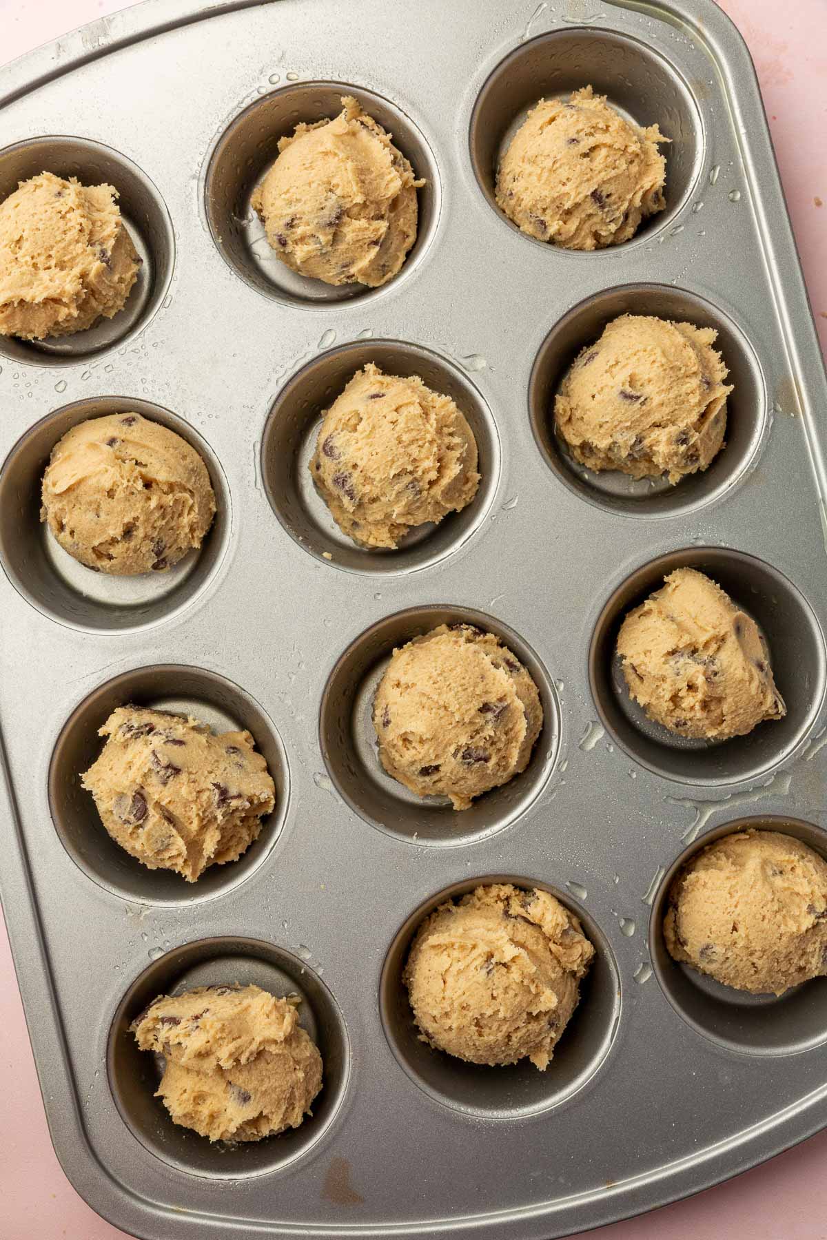 A muffin tin that has scoops of gluten-free chocolate chip cookie dough in the center of each muffin well before baking in the oven.