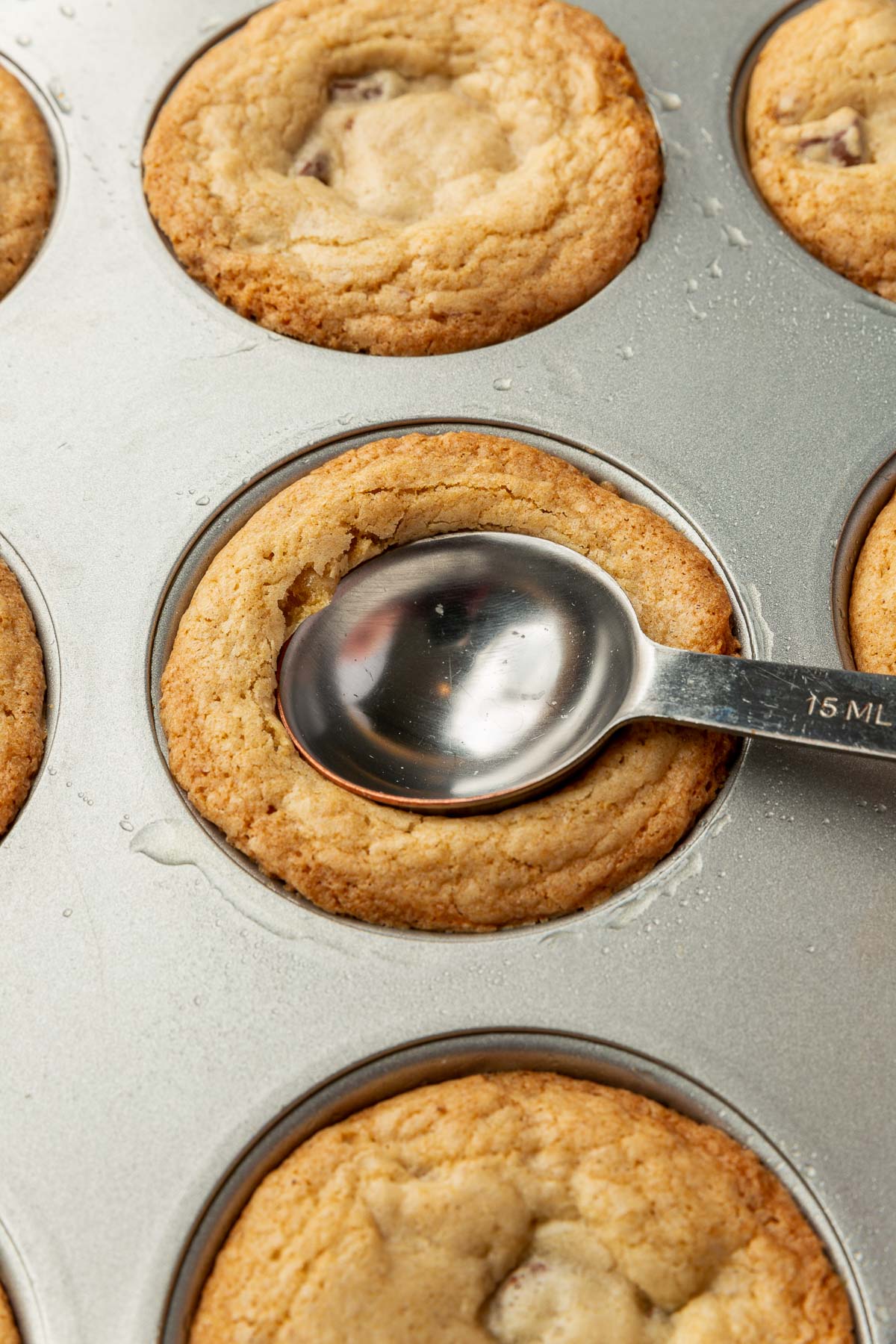 A tablespoon-sized measuring spoon pressing down in a chocolate chip cookie cup in a muffin tin to make an indentation after baking in the oven.