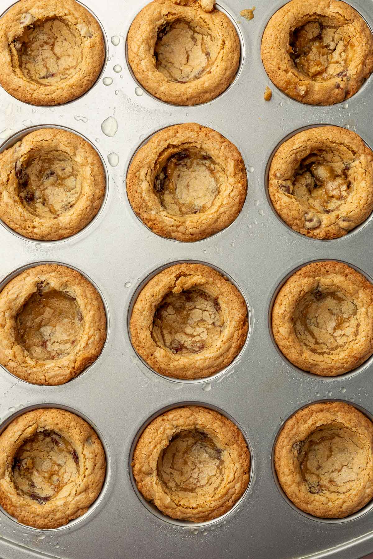 An overhead view of muffin-shaped cookie cups in a muffin tin that have been pressed down in the center of each cookie cup to make an indentation for frosting.