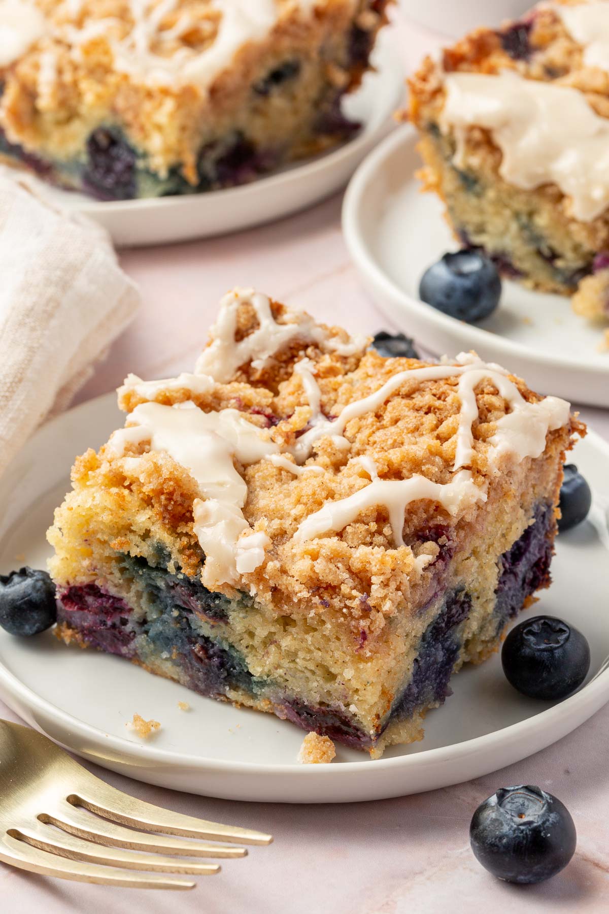 A slice of gluten-free blueberry coffee cake with streusel and glaze on a dessert plate with two more slices behind it.