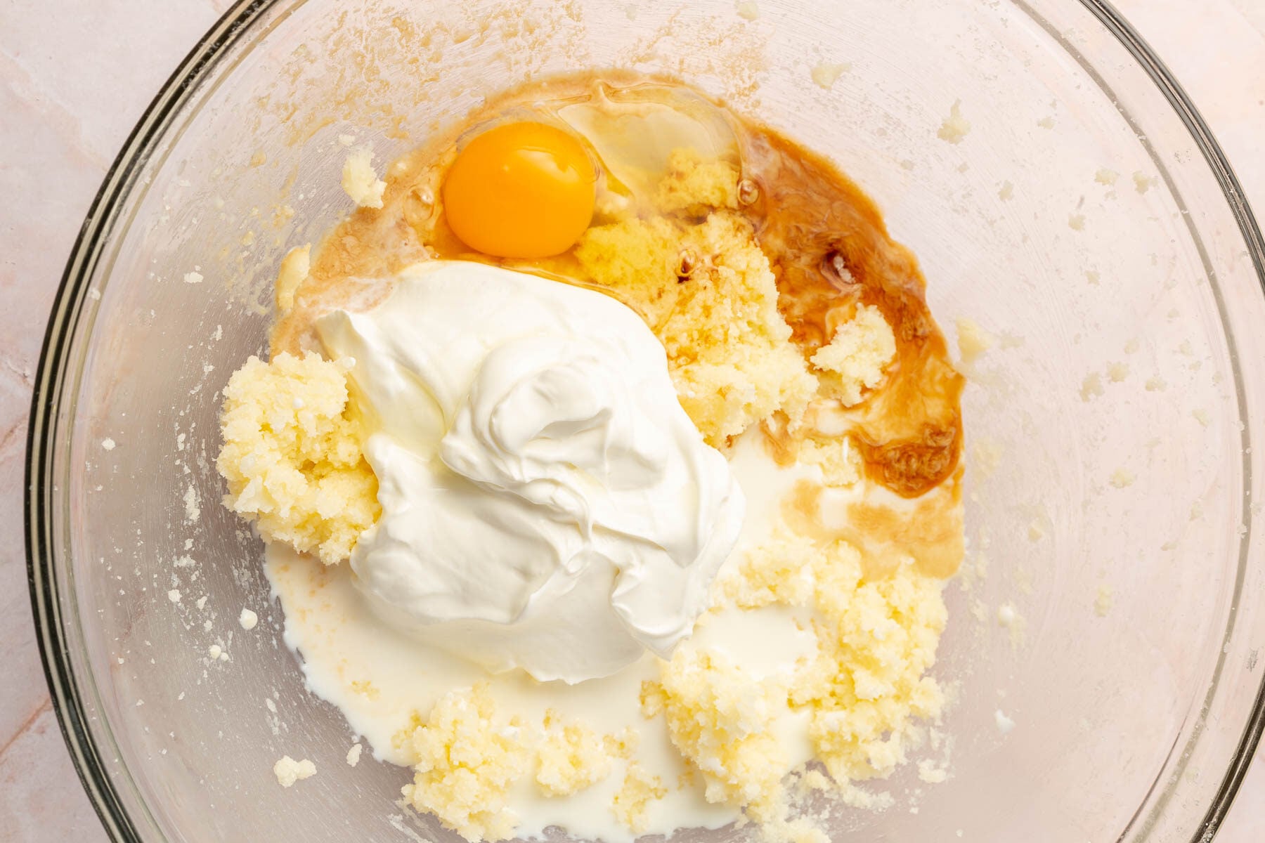 A glass mixing bowl with creamed butter and sugar topped with sour cream, milk, egg and vanilla before mixing together.