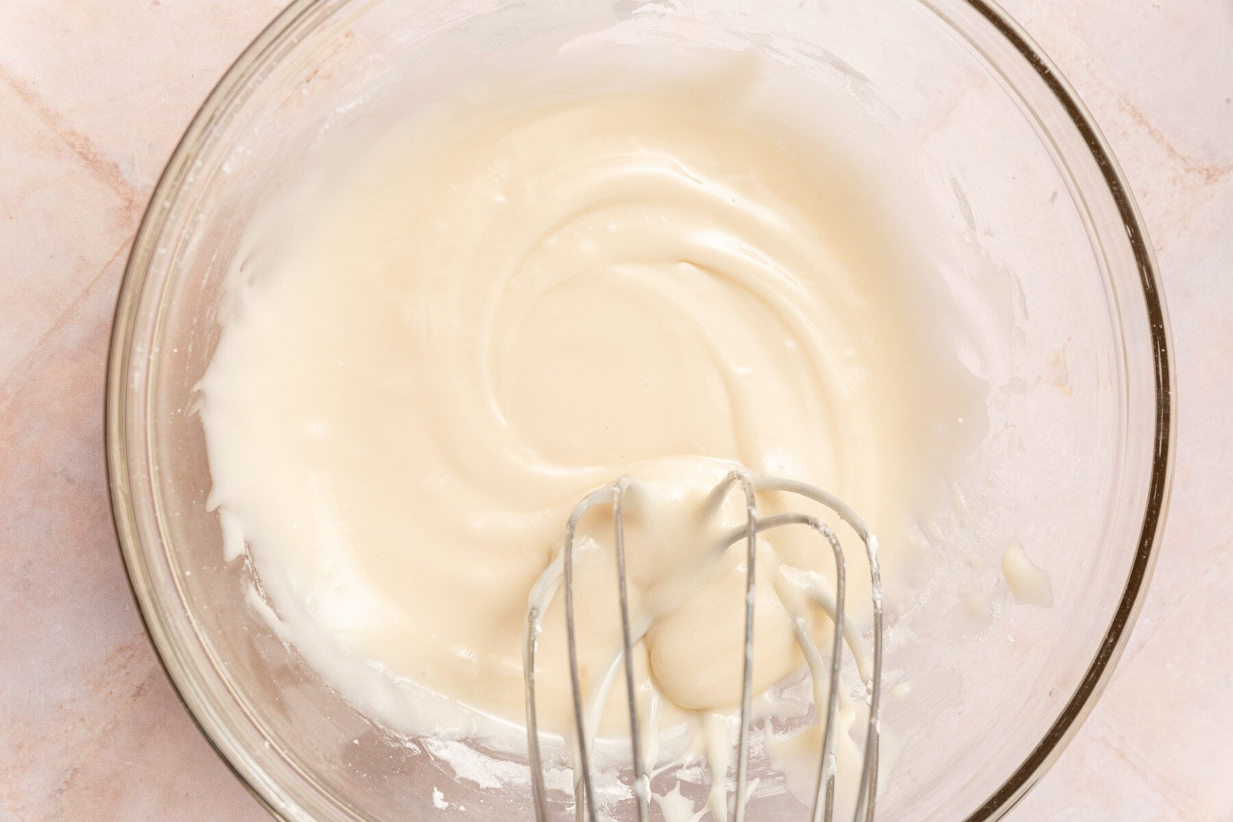 A glass mixing bowl filled with a vanilla glaze being mixed with a wire whisk.