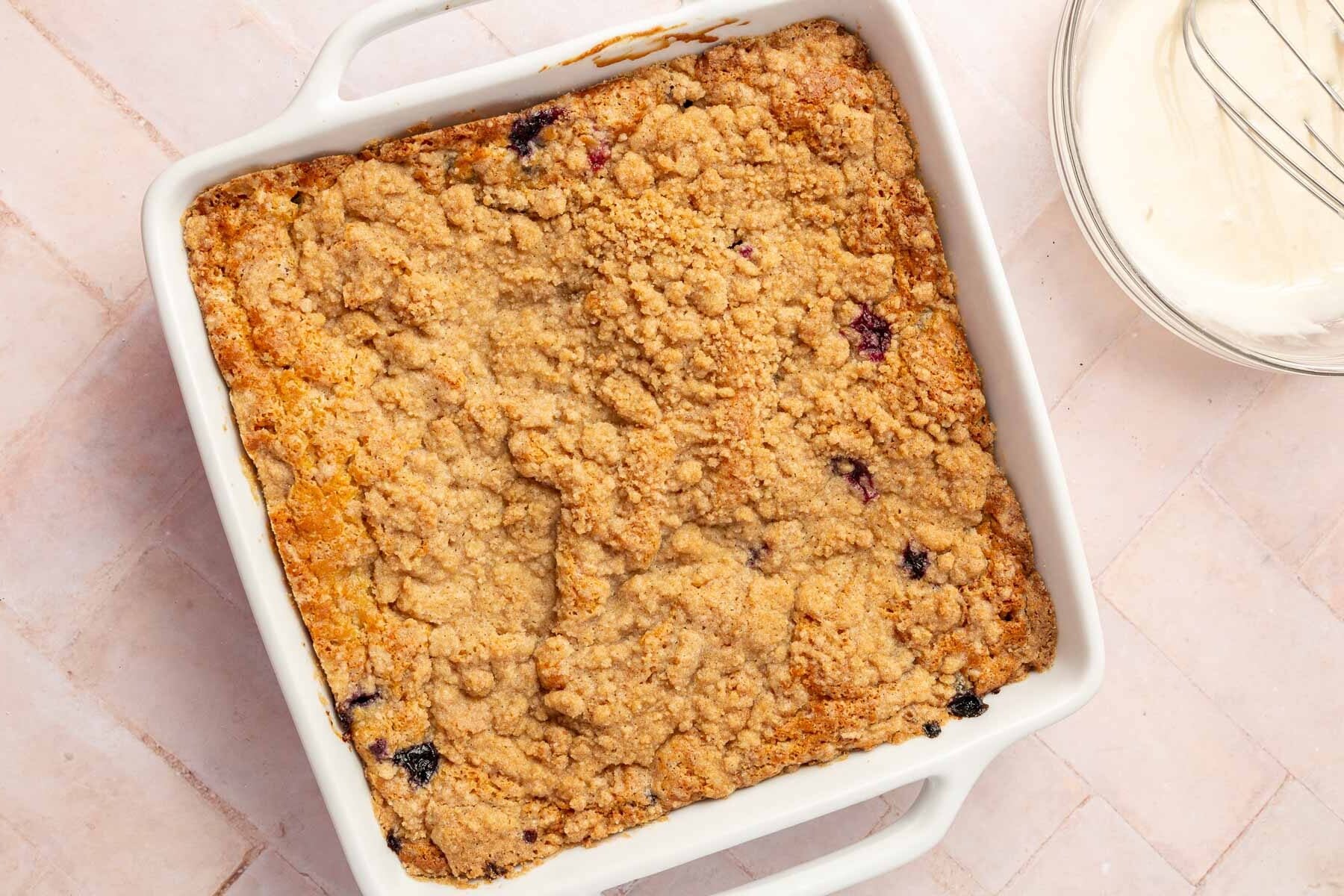 An overhead photo of a square ceramic baking dish filled with a gluten free blueberry coffee cake just out of the oven with a bowl of glaze off to the side.