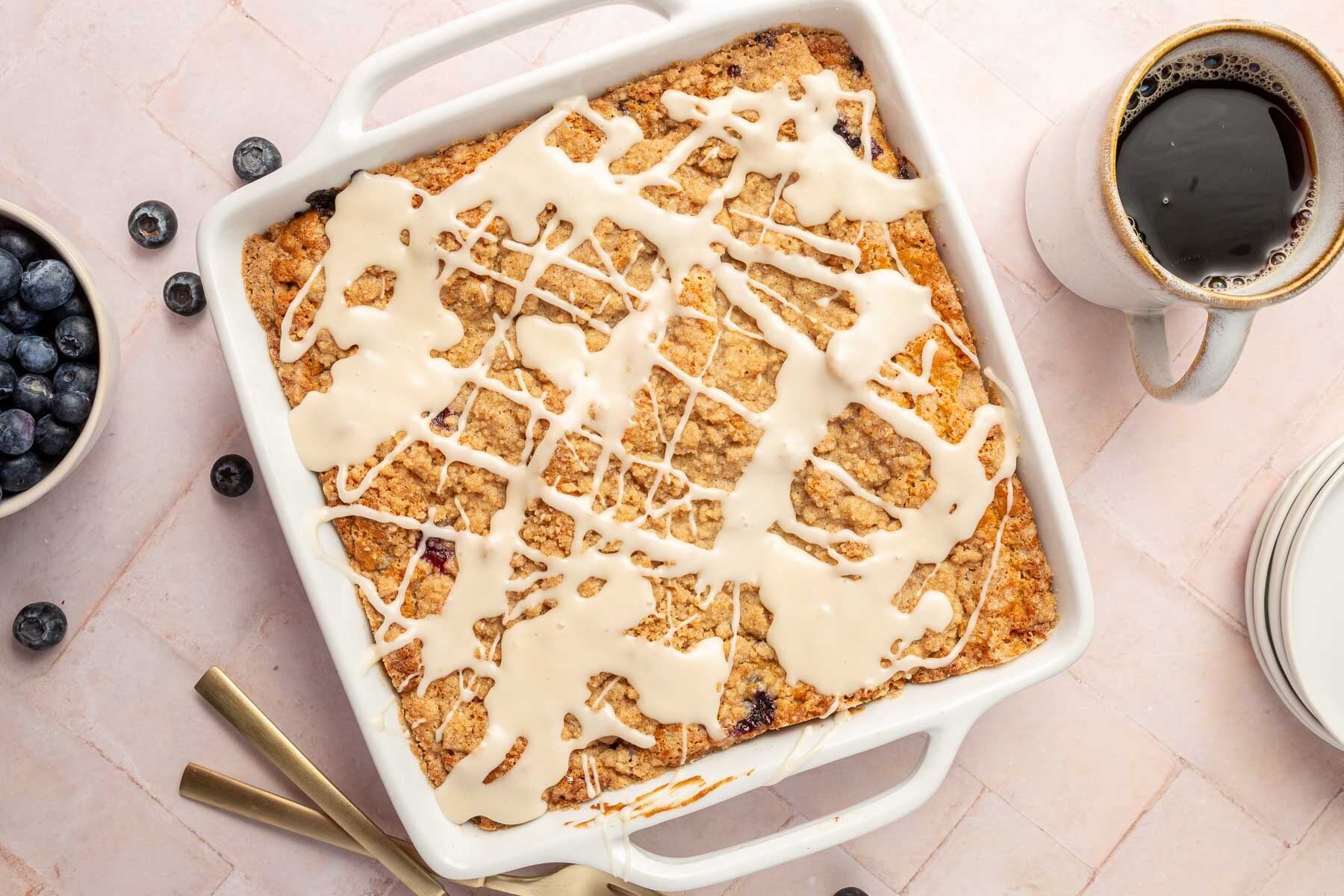 A square baking dish filled with a blueberry coffee cake with streusel and drizzled glaze on top with a cup of coffee and a bowl of fresh blueberries off to the side.
