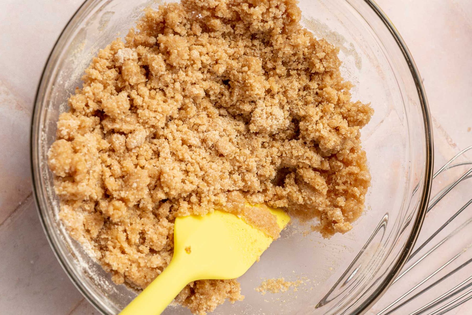 A glass mixing bowl with gluten free streusel topping and a yellow spatula in it.
