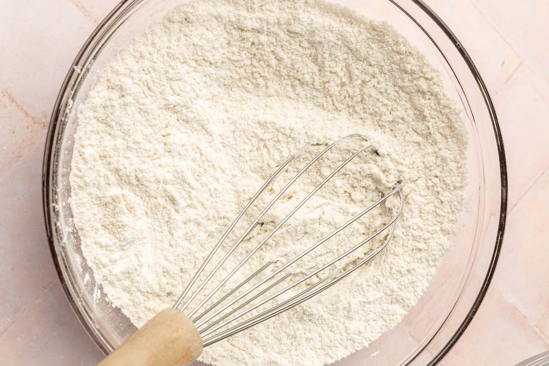 A glass mixing bowl of gluten-free flour, baking soda, baking powder and salt whisked together with a wire whisk.