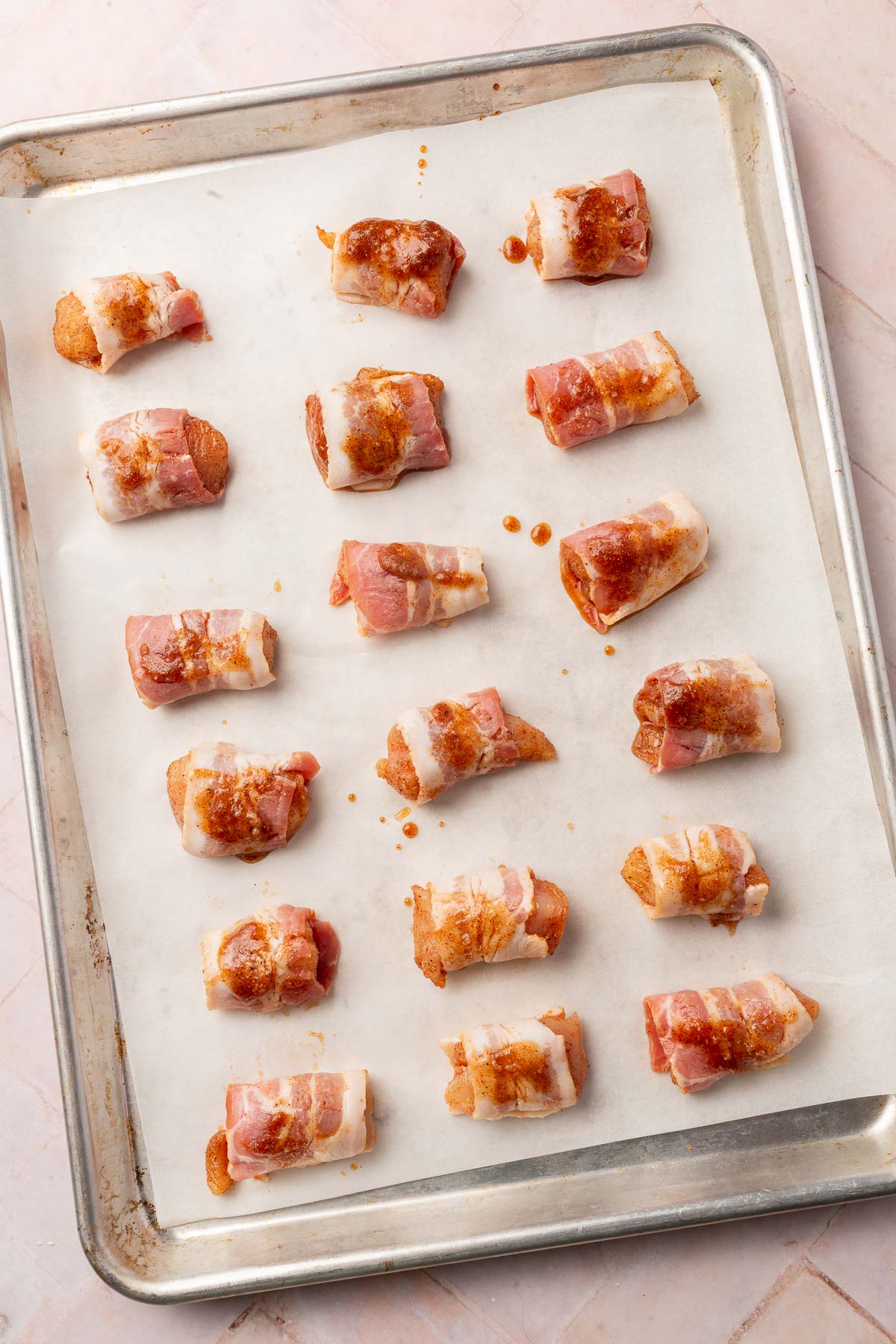 An overhead view of a parchment lined baking sheet with rows of bacon wrapped chicken bites before putting in the oven.