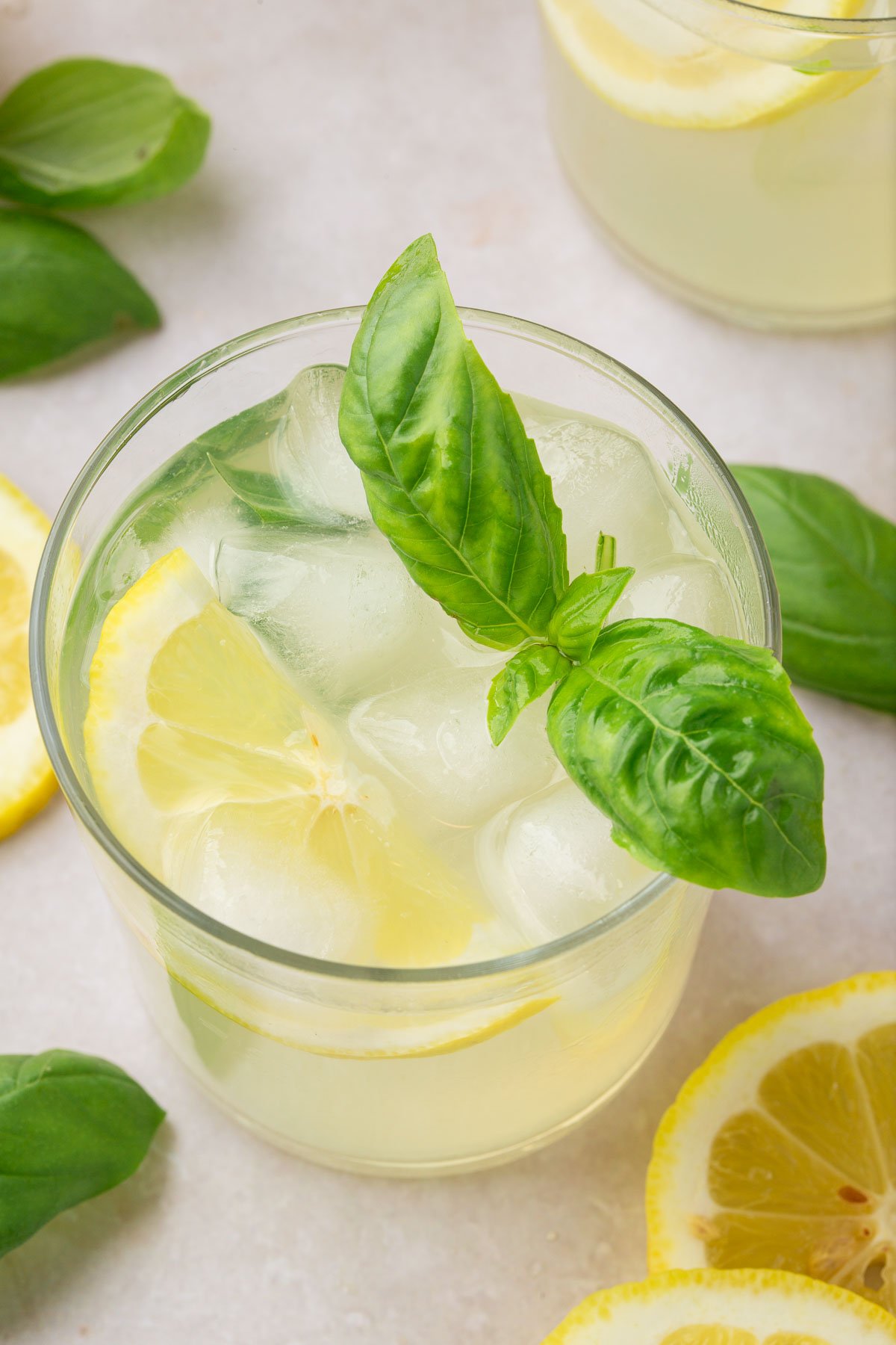 A glass of basil lemonade filled with ice and topped with a slice of lemon and a few basil leaves with additional lemon slices and fresh basil surrounding the glass.