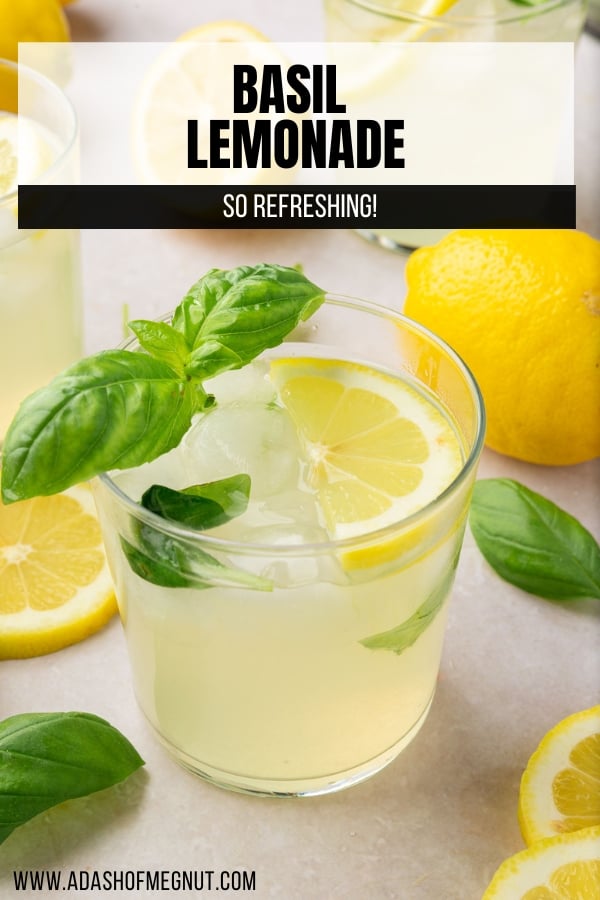 A glass of basil lemonade filled with ice and topped with a slice of lemon and a few basil leaves with additional lemon slices and fresh basil surrounding the glass.