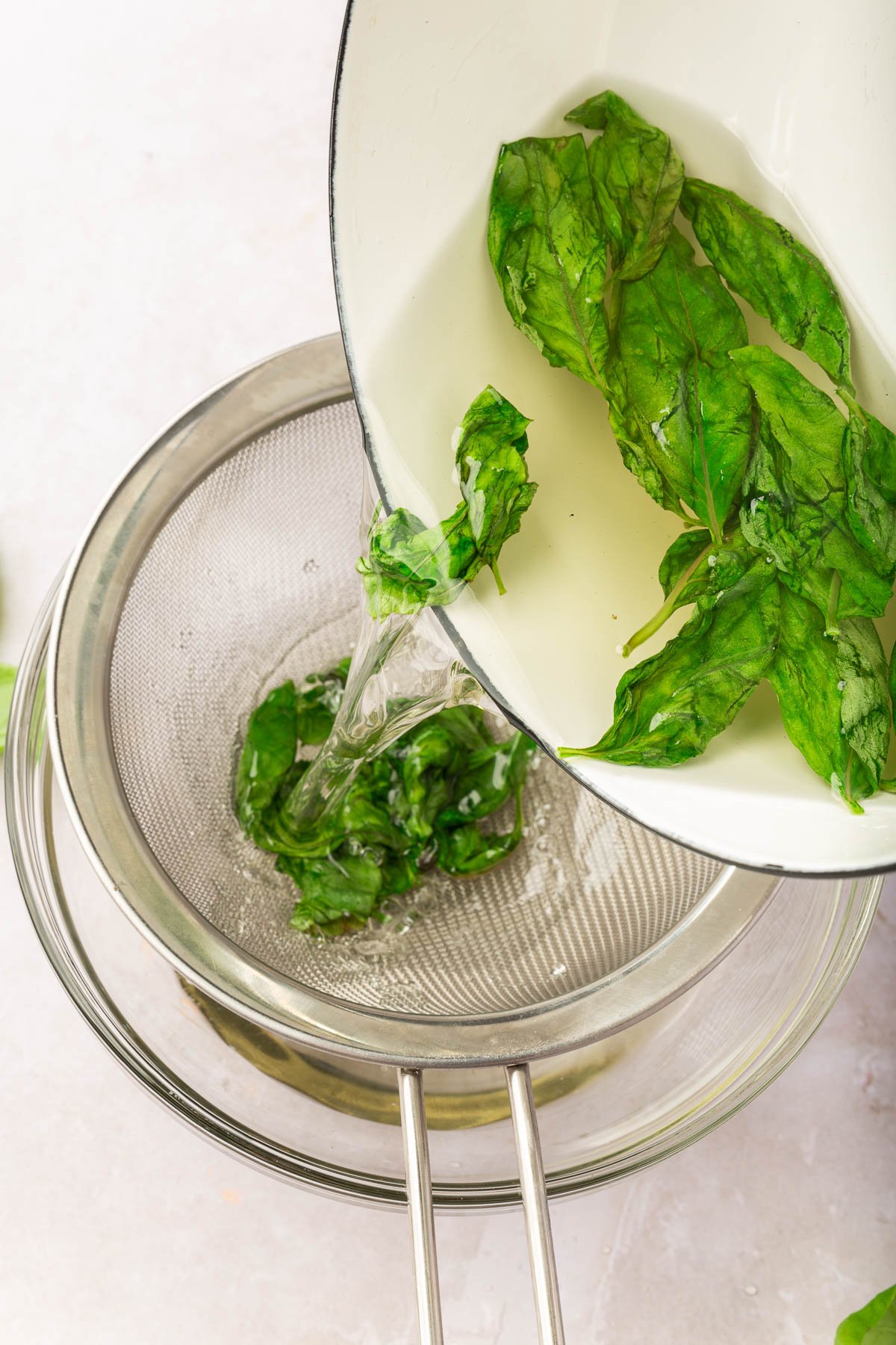 A saucepan pouring fresh basil leaves and simply syrup into a wire mesh sieve to strain out the basil leaves.