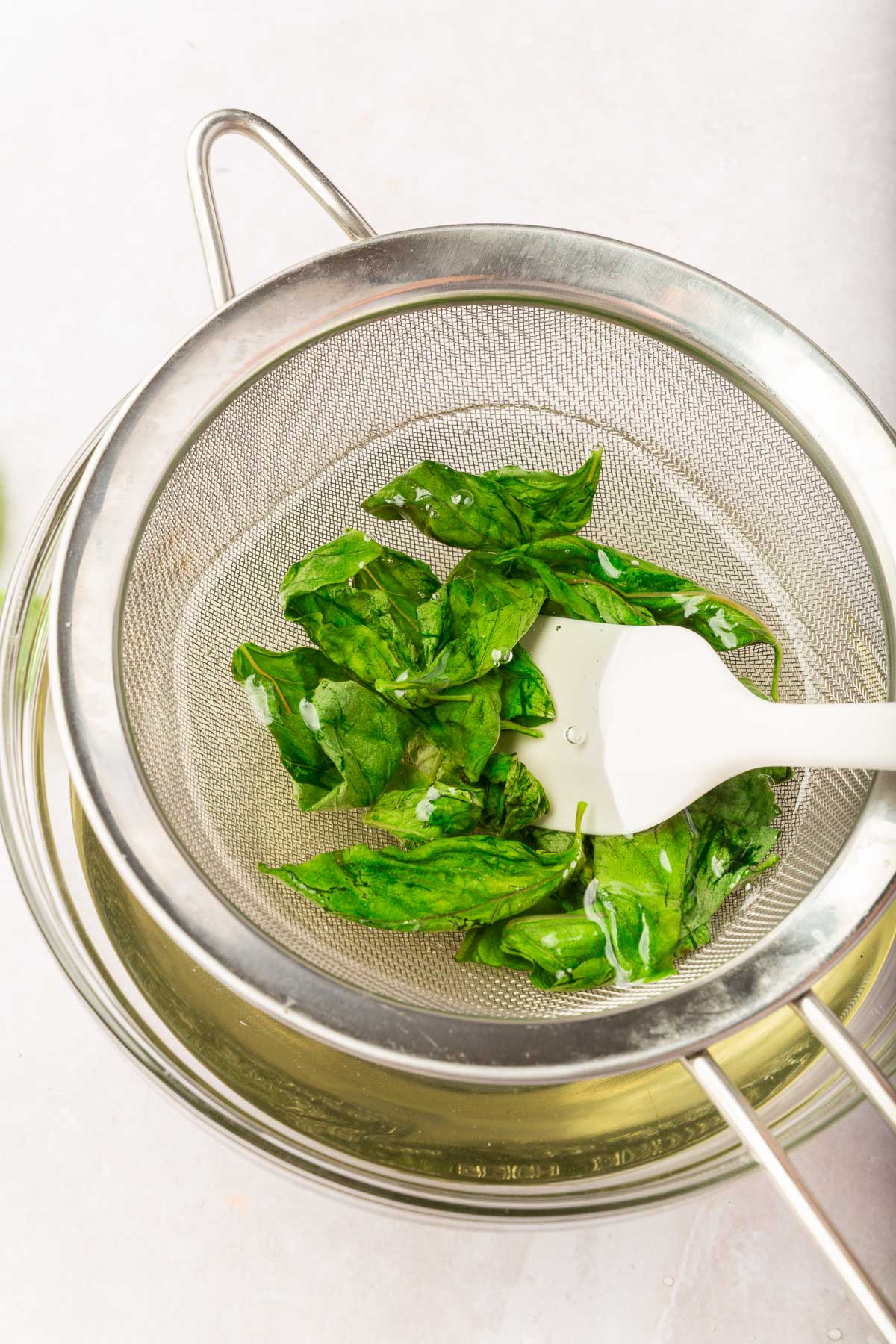 Fresh basil leaves being pressed down into a wire mesh sieve to strain it from simple syrup.
