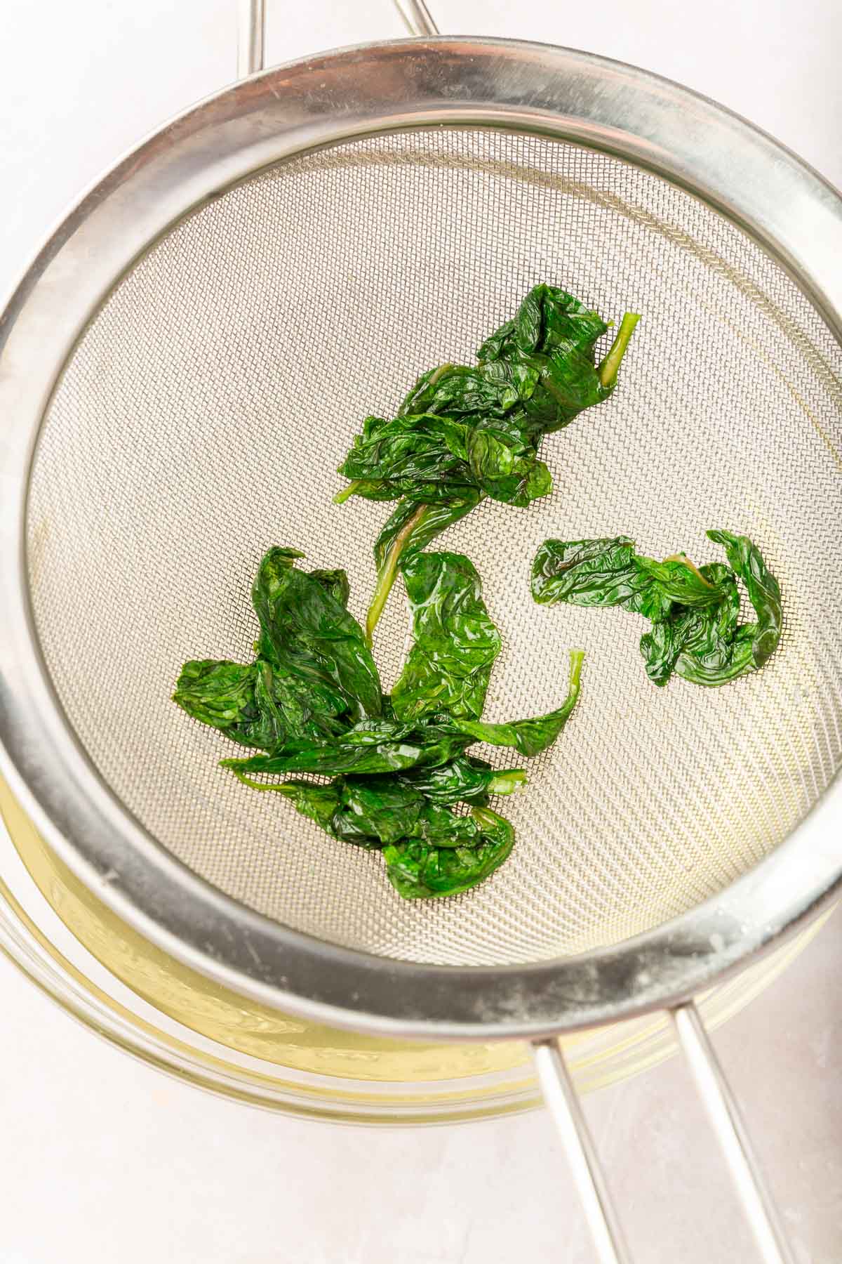 Wilted basil being strained from a simple syrup with a mesh wire sieve over a bowl of simple syrup.