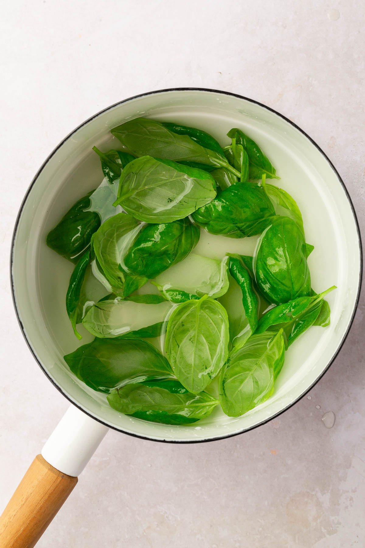 An overhead view of water, granulated sugar and basil leaves in a white saucepan to make basil simple syrup.