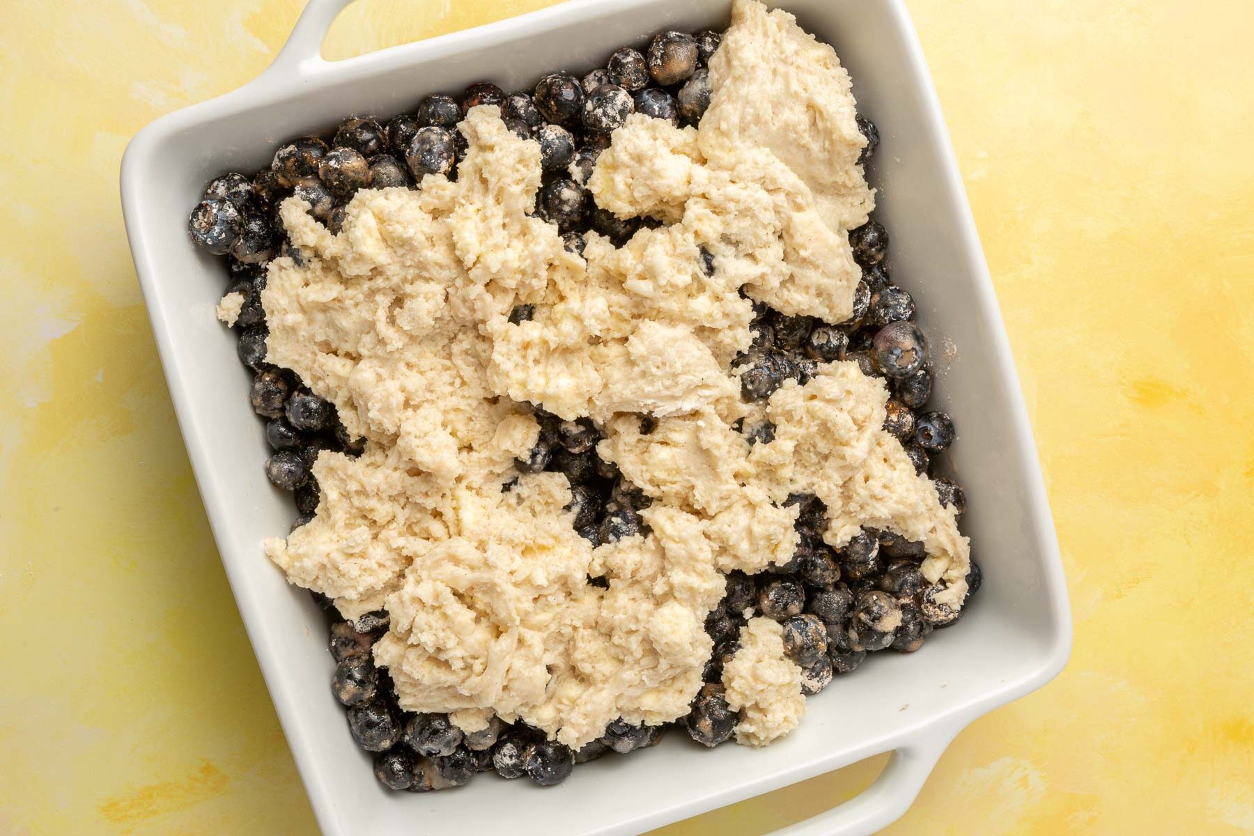 A square baking dish filled with blueberries topped with raw gluten free biscuit dough before baking in the oven.