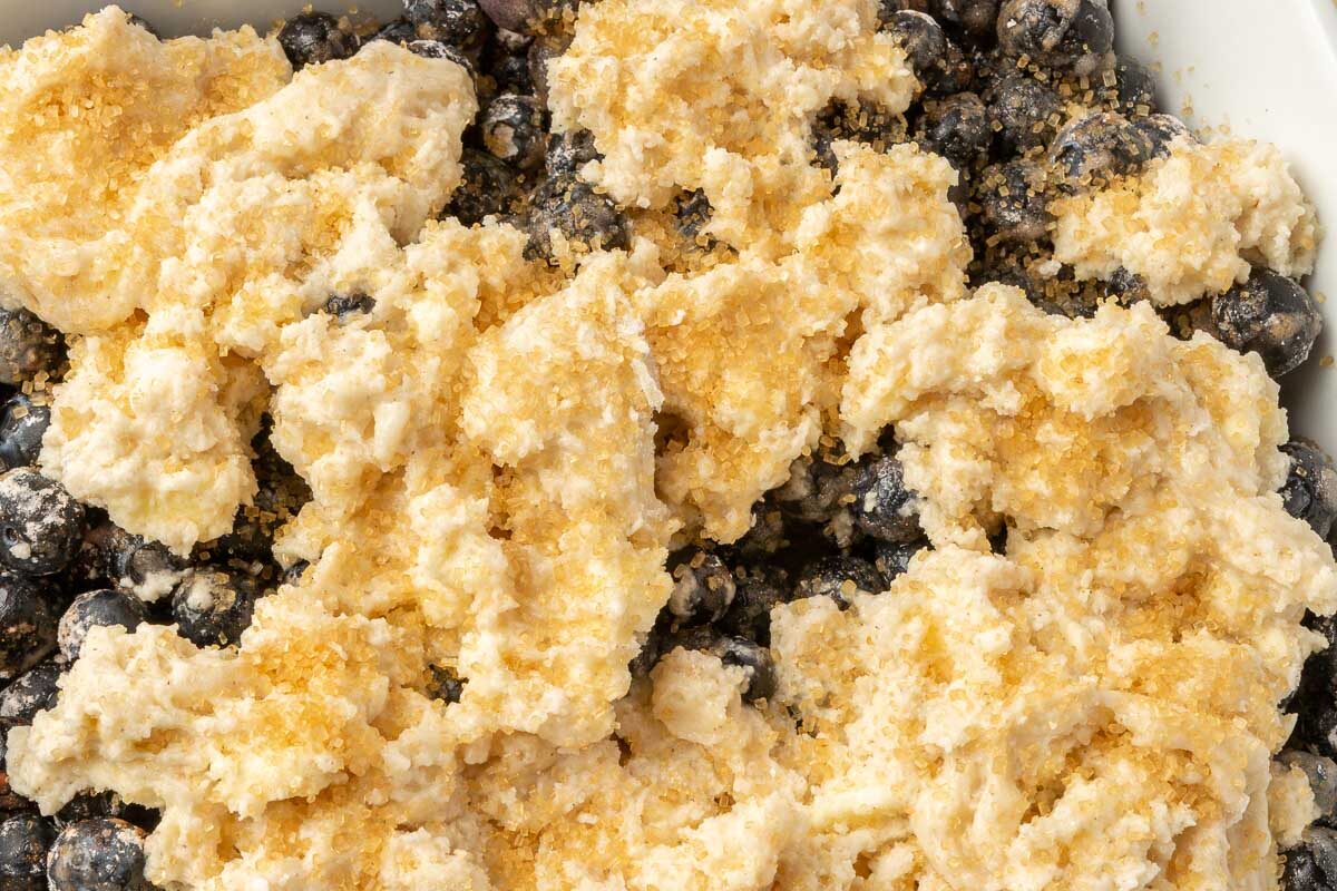 A square baking dish filled with blueberries topped with raw gluten free biscuit dough and turbinado sugar before baking in the oven.