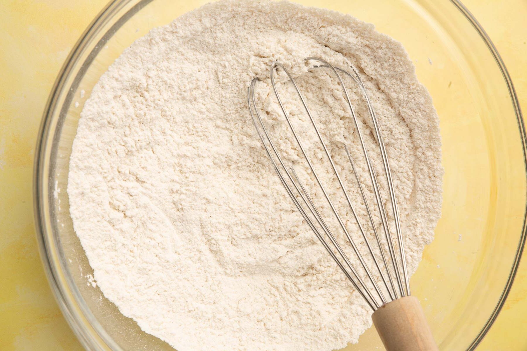 A glass mixing bowl with granulated sugar, gluten-free flour, baking soda, baking powder and salt mixed together with a wire whisk.