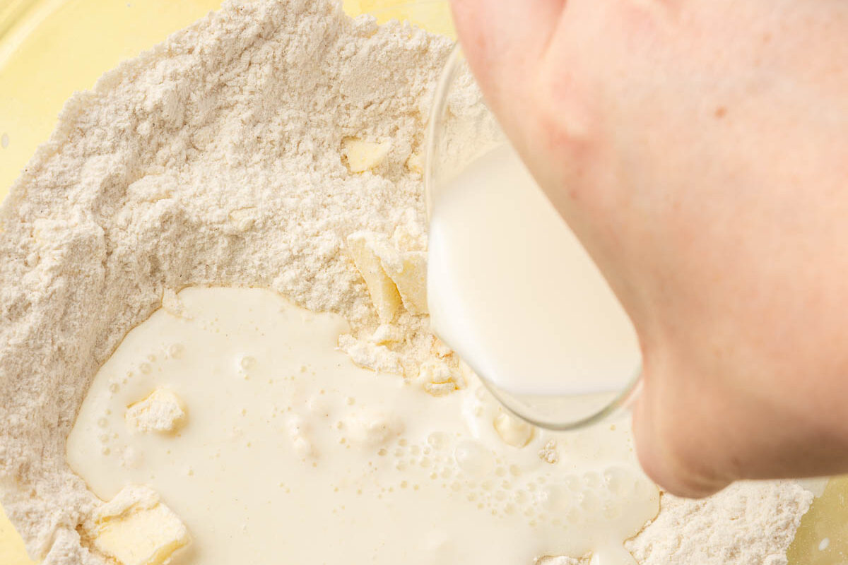 A hand pouring buttermilk into a glass bowl of gluten-free flour and cubes of butter.