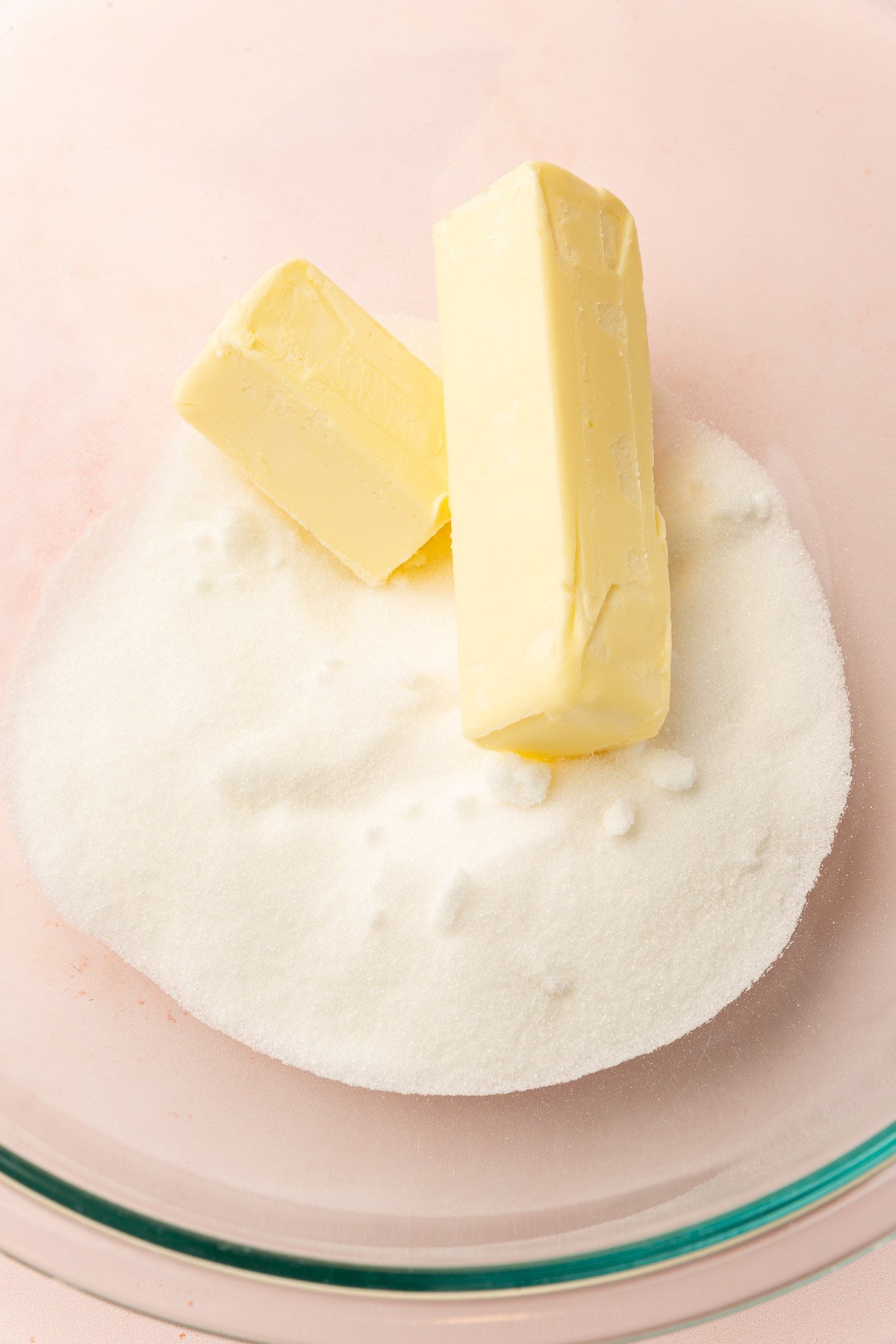 A glass mixing bowl with granulated sugar and two sticks of unsalted butter in it before mixing together.