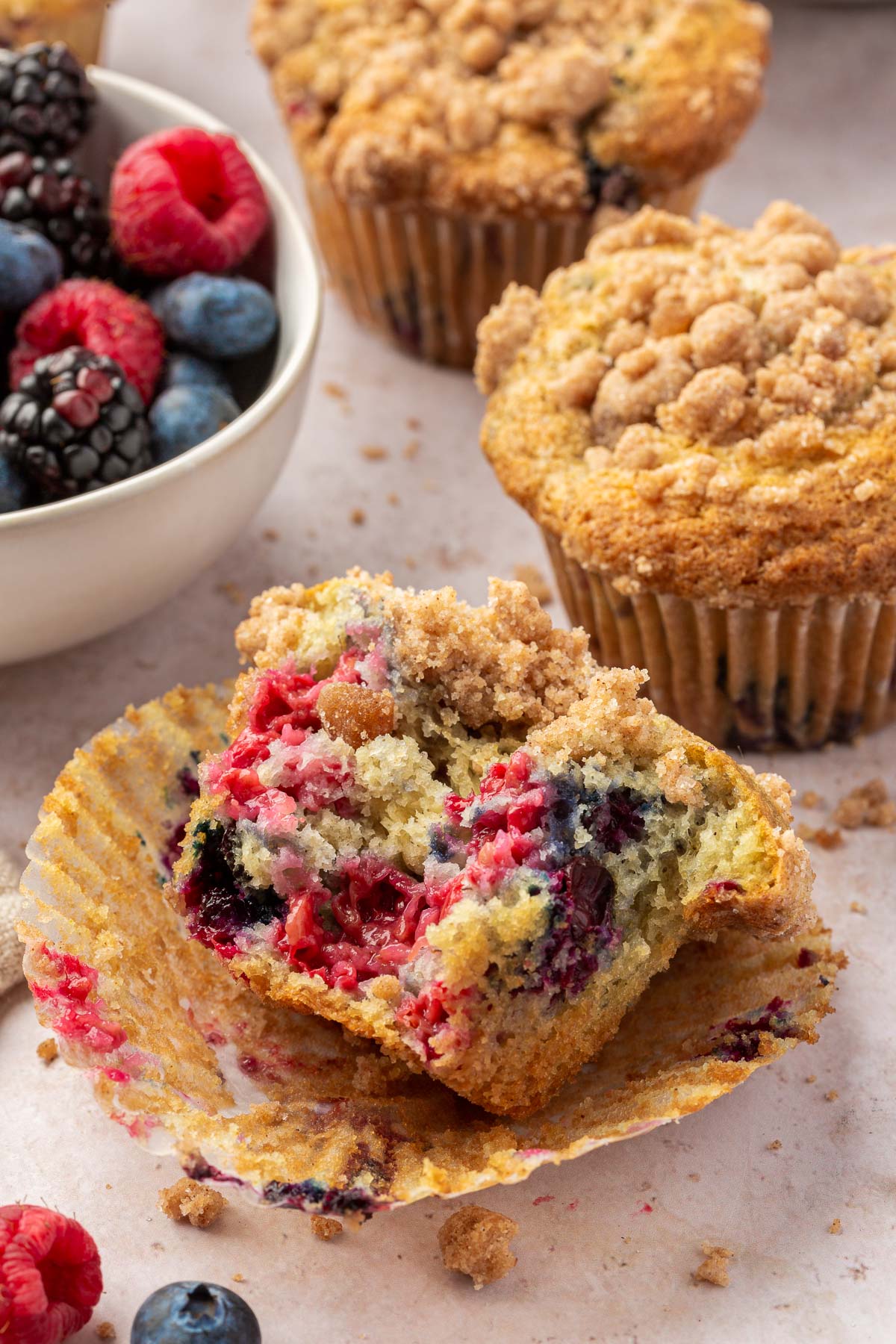 Half of a gf triple berry muffin that has been bitten into and is laying on the muffin liner with additional muffins and streusel crumbs in the background.