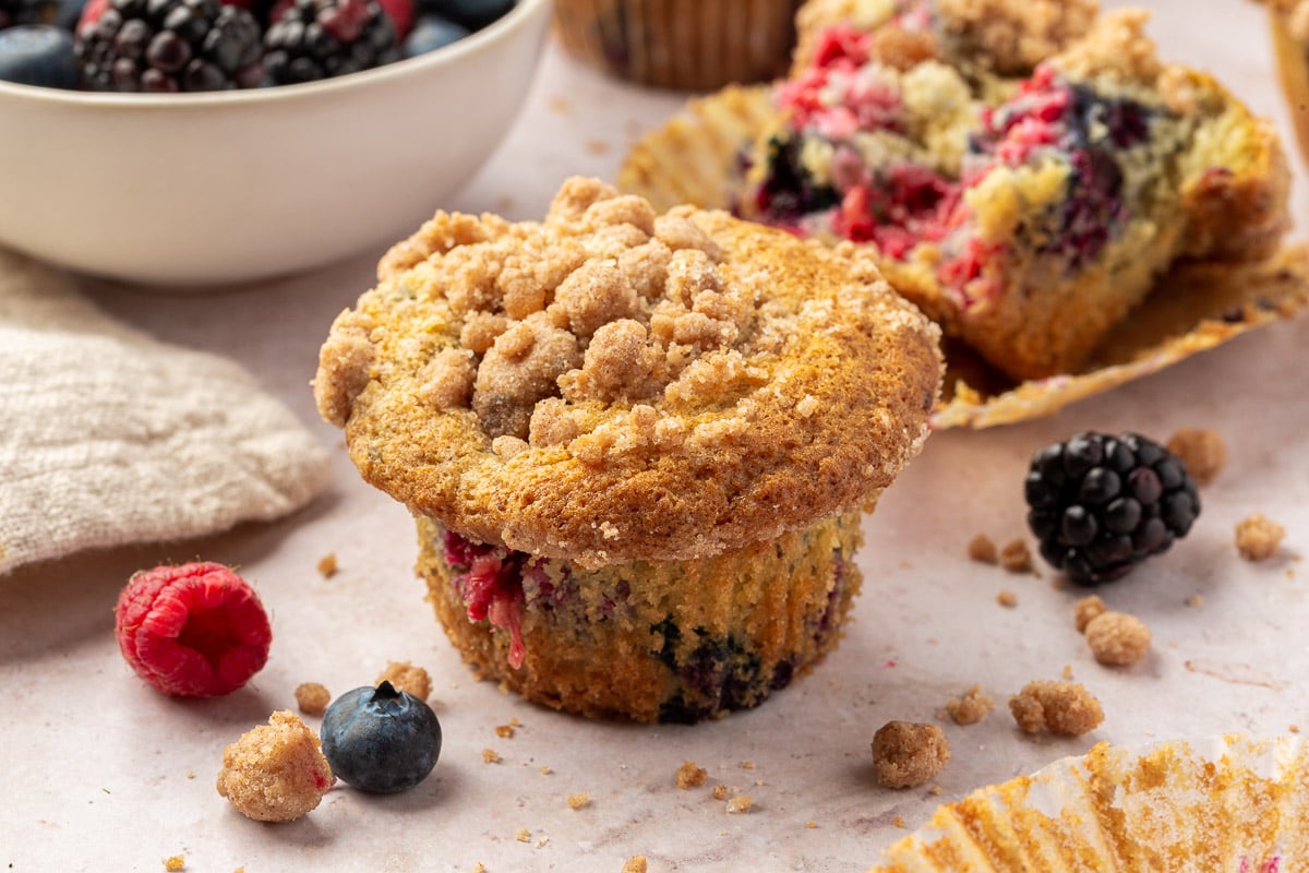 A gluten-free mixed berry muffin with streusel topping surrounded by additional blueberries, raspberries and blackberries.