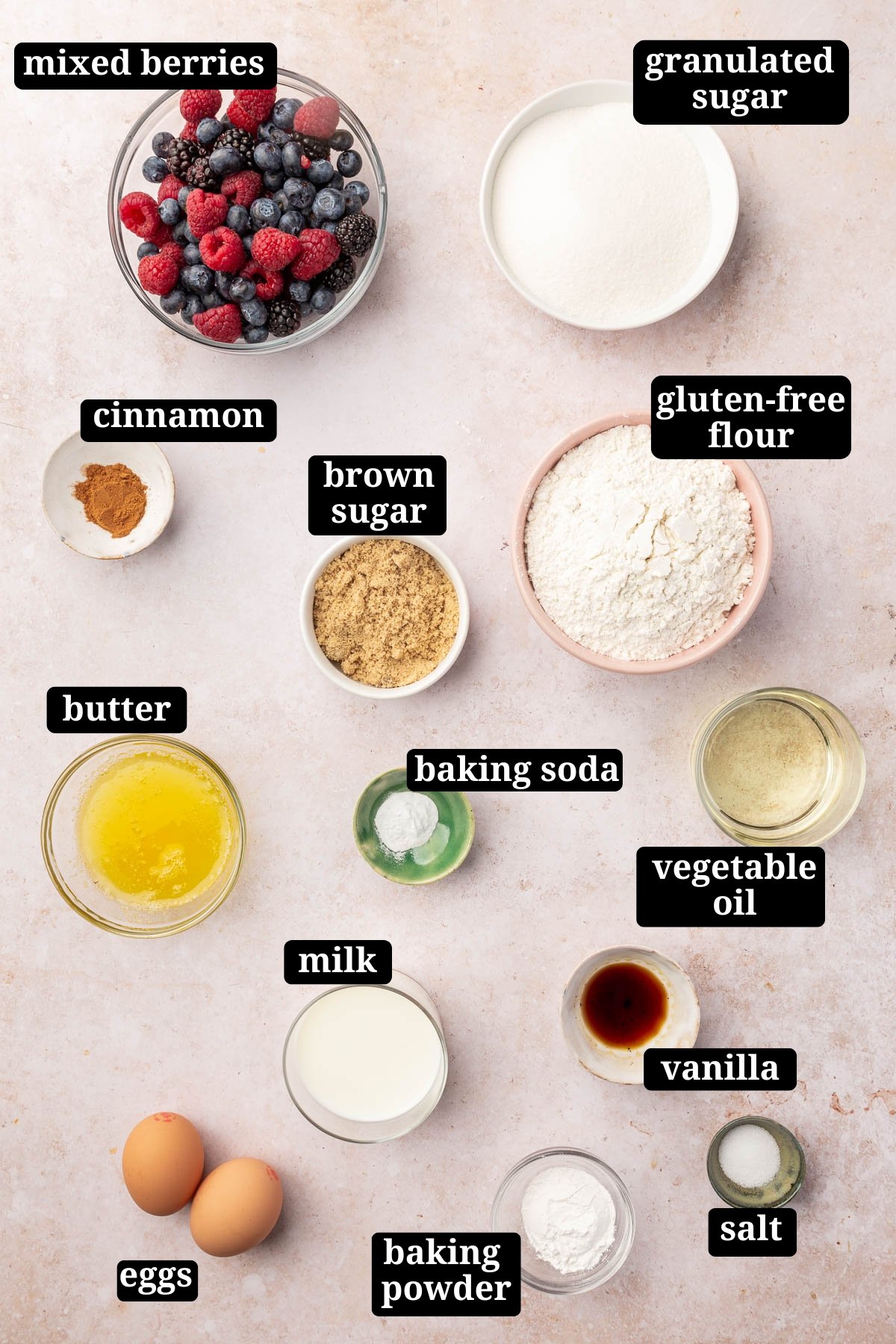 Ingredients in small bowls to make gluten-free mixed berry muffins with streusel, including mixed berries, granulated sugar, cinnamon, brown sugar, gluten-free flour, butter, baking soda, vegetable oil, milk, vanilla, salt, baking powder and eggs with text overlays over each ingredient.