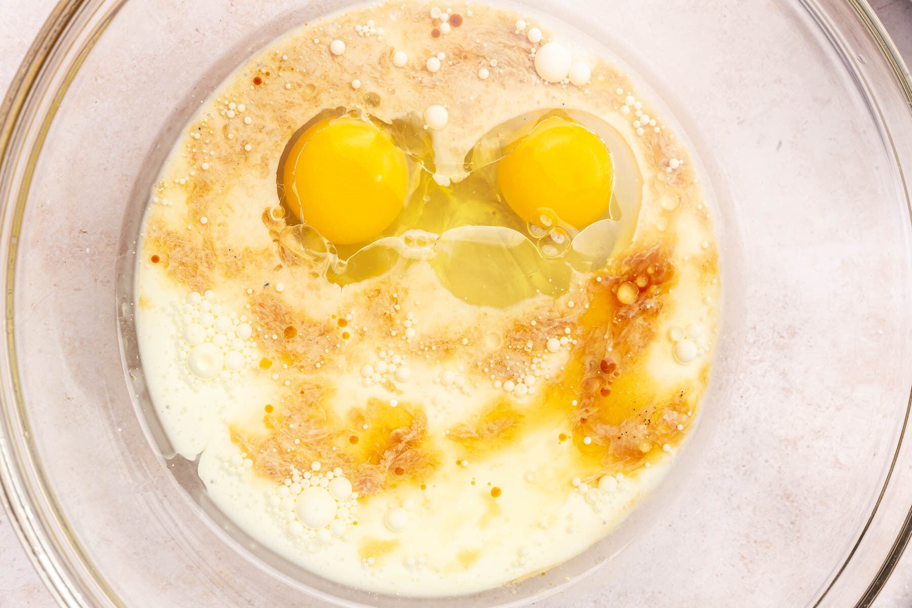 A glass mixing bowl with eggs, milk, oil, and vanilla in it before whisking together.