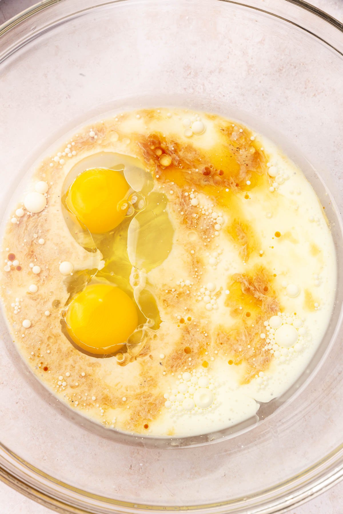 A glass mixing bowl with eggs, milk, oil, and vanilla in it before whisking together.