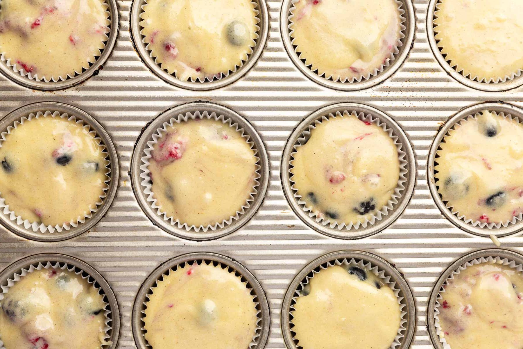 An overhead view of a muffin tin filled with 12 muffin liners of gluten-free mixed berry muffin batter before baking in the oven.
