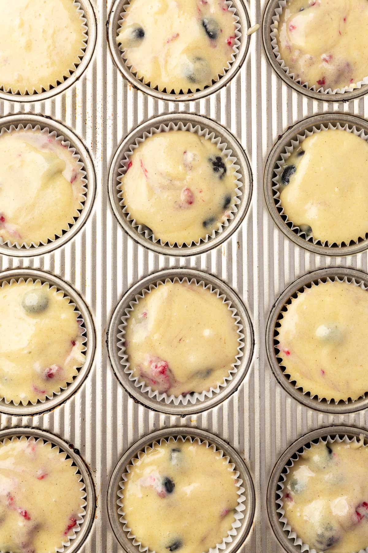 An overhead view of a muffin tin filled with 12 muffin liners of gluten-free mixed berry muffin batter before baking in the oven.